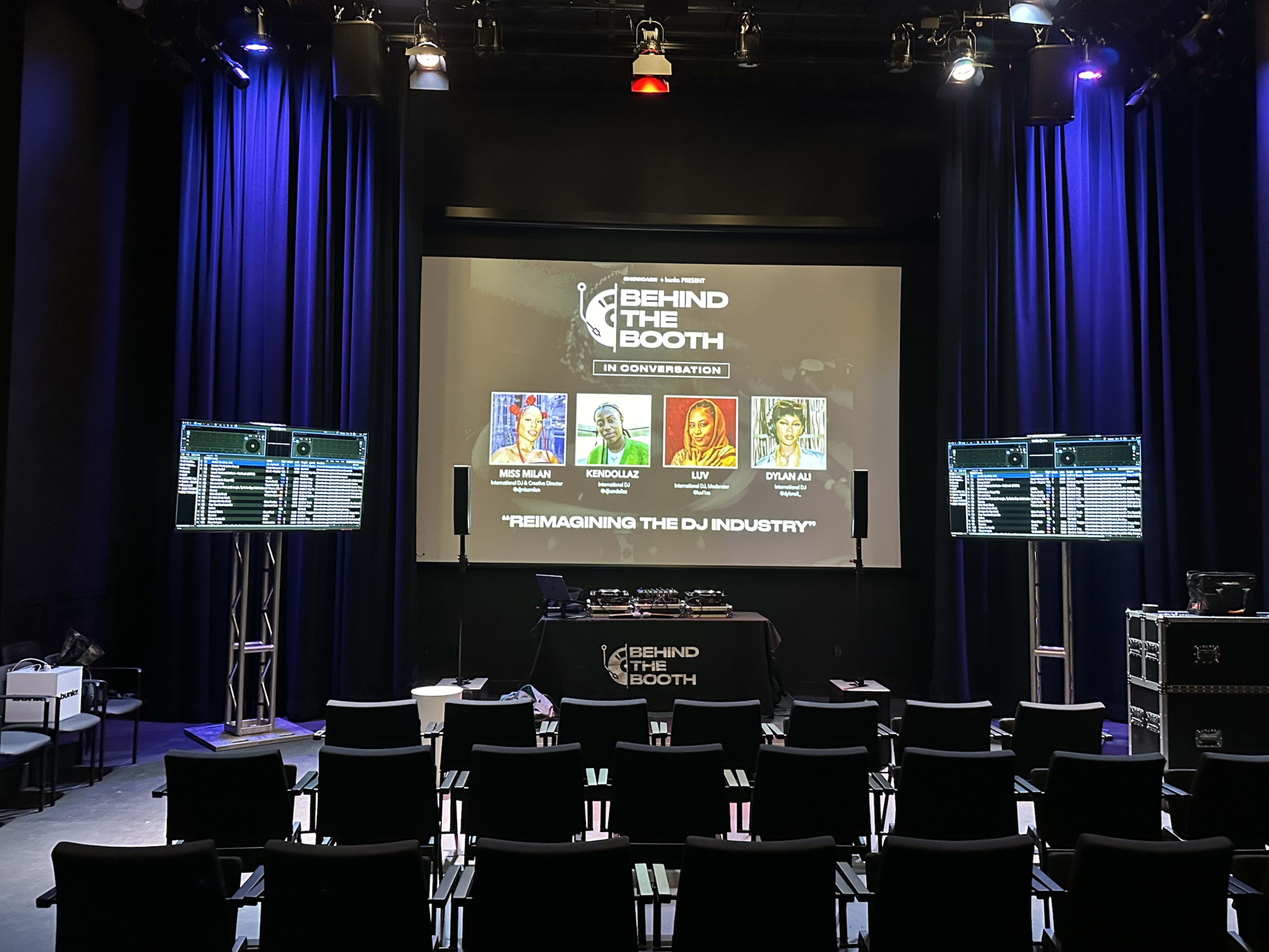 A stage setup for an event titled "Behind the Booth" with a large screen displaying the event name and four speakers' portraits, alongside equipment and chairs arranged in a theater-style seating.