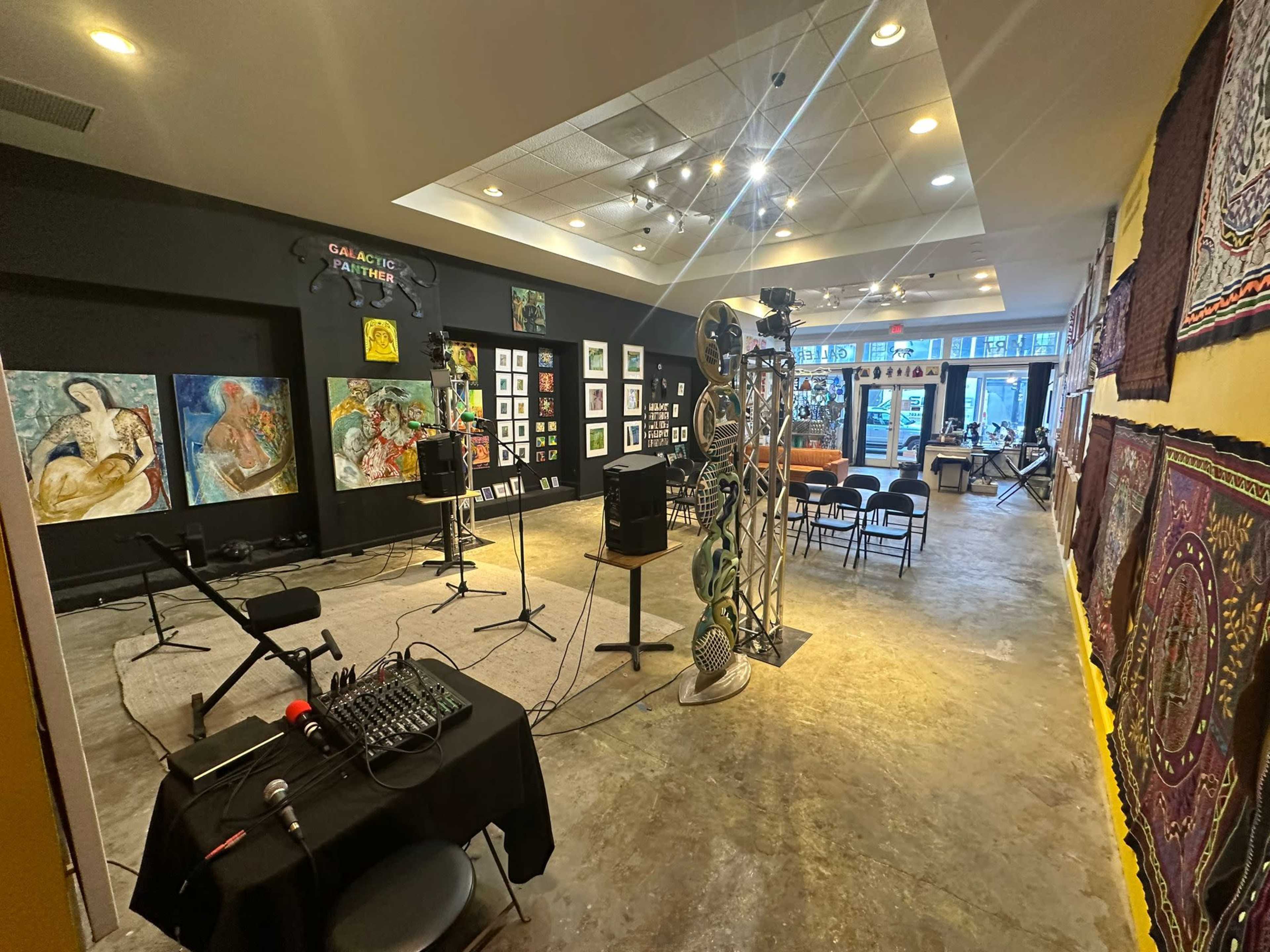 The image shows an art gallery space set up for a performance, with paintings on the walls, a sound mixer on a table, and chairs arranged for an audience.