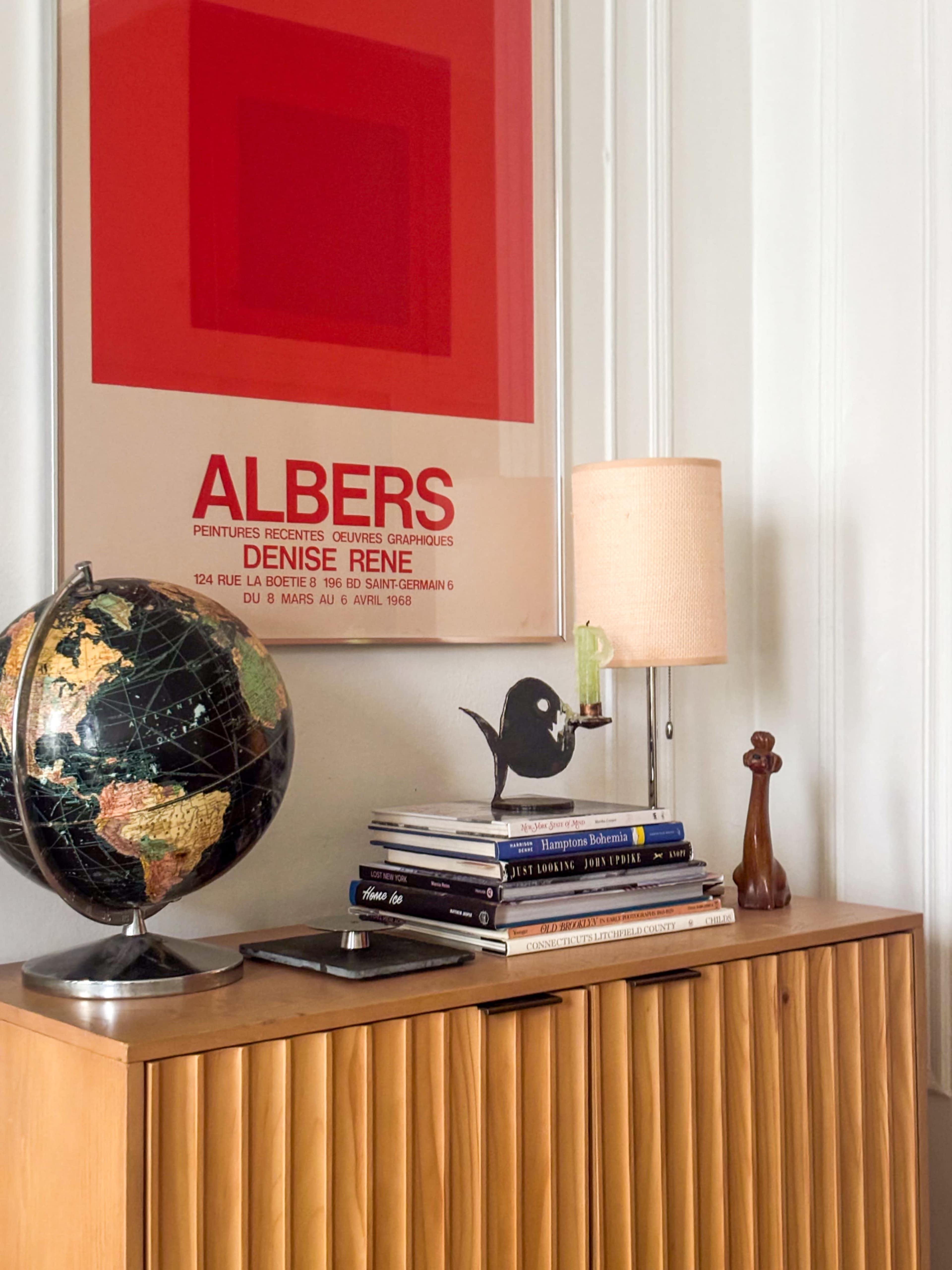 The image shows a wooden cabinet topped with a globe, a lamp, and stacks of books, alongside a framed art poster featuring bold red and white colors.