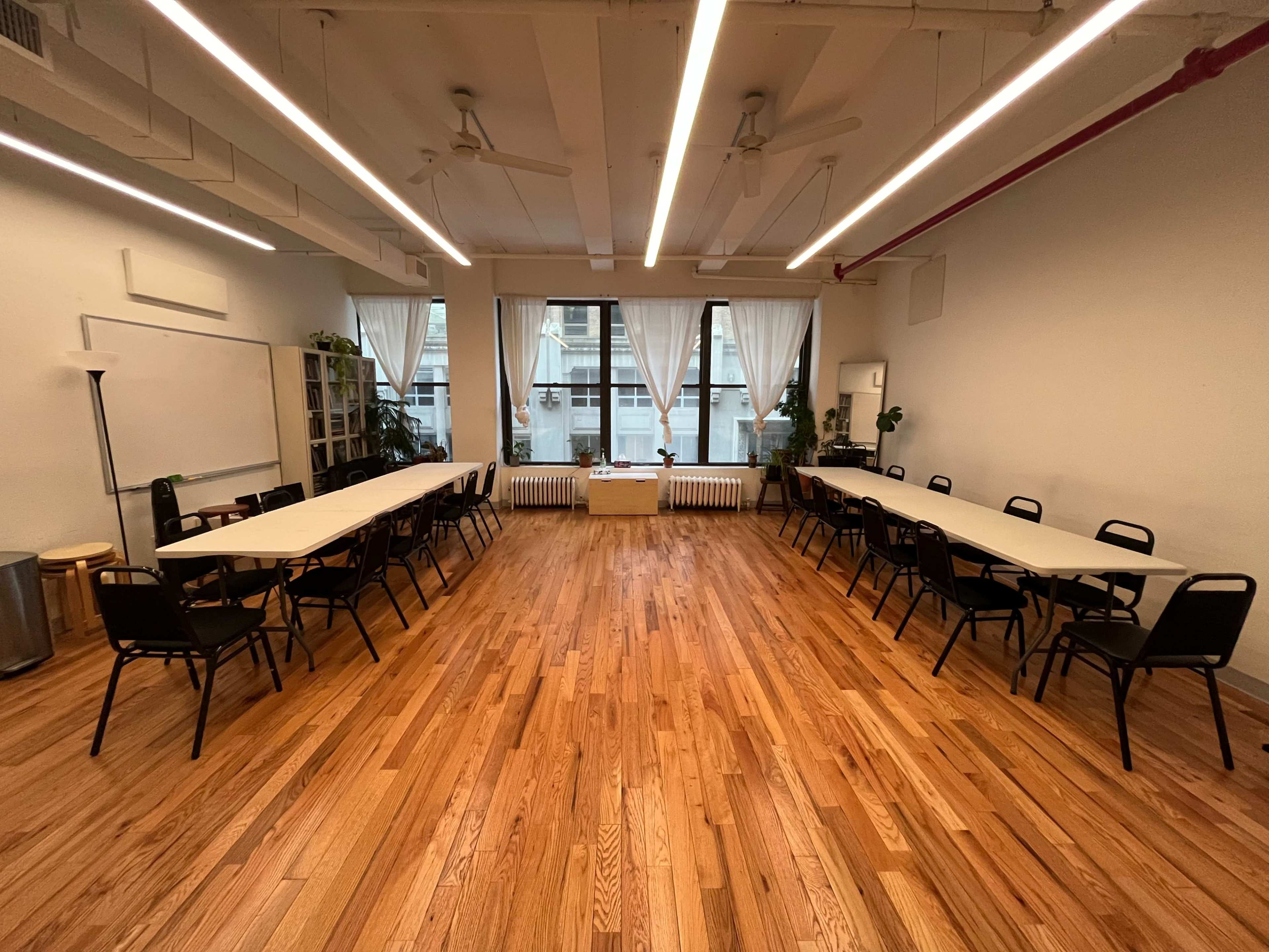 A spacious meeting room with two long tables arranged in a U-shape, surrounded by black chairs and large windows with natural light.