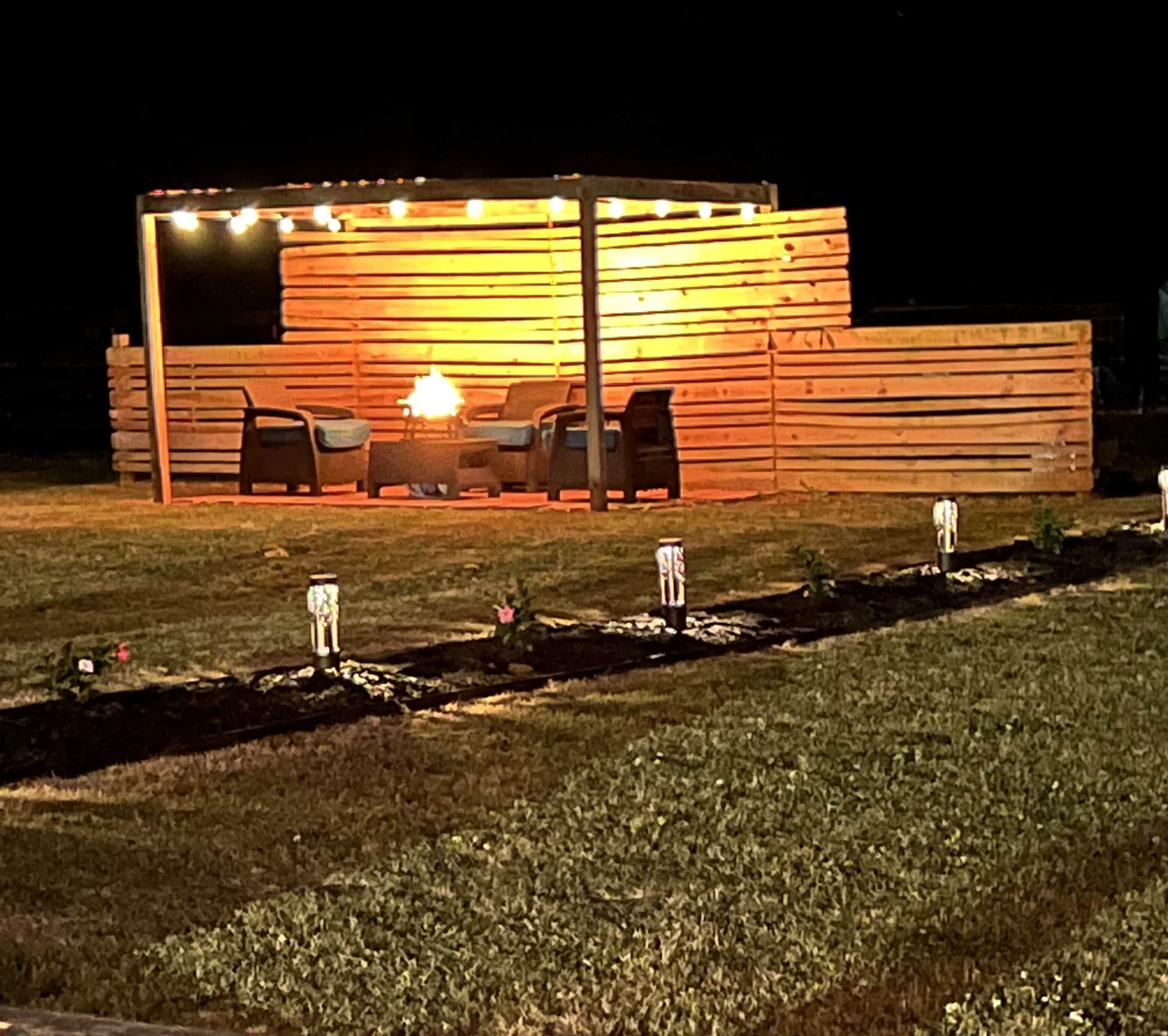 A lit outdoor seating area features a fire pit surrounded by chairs and a wooden structure with lights, set against a grassy landscape.