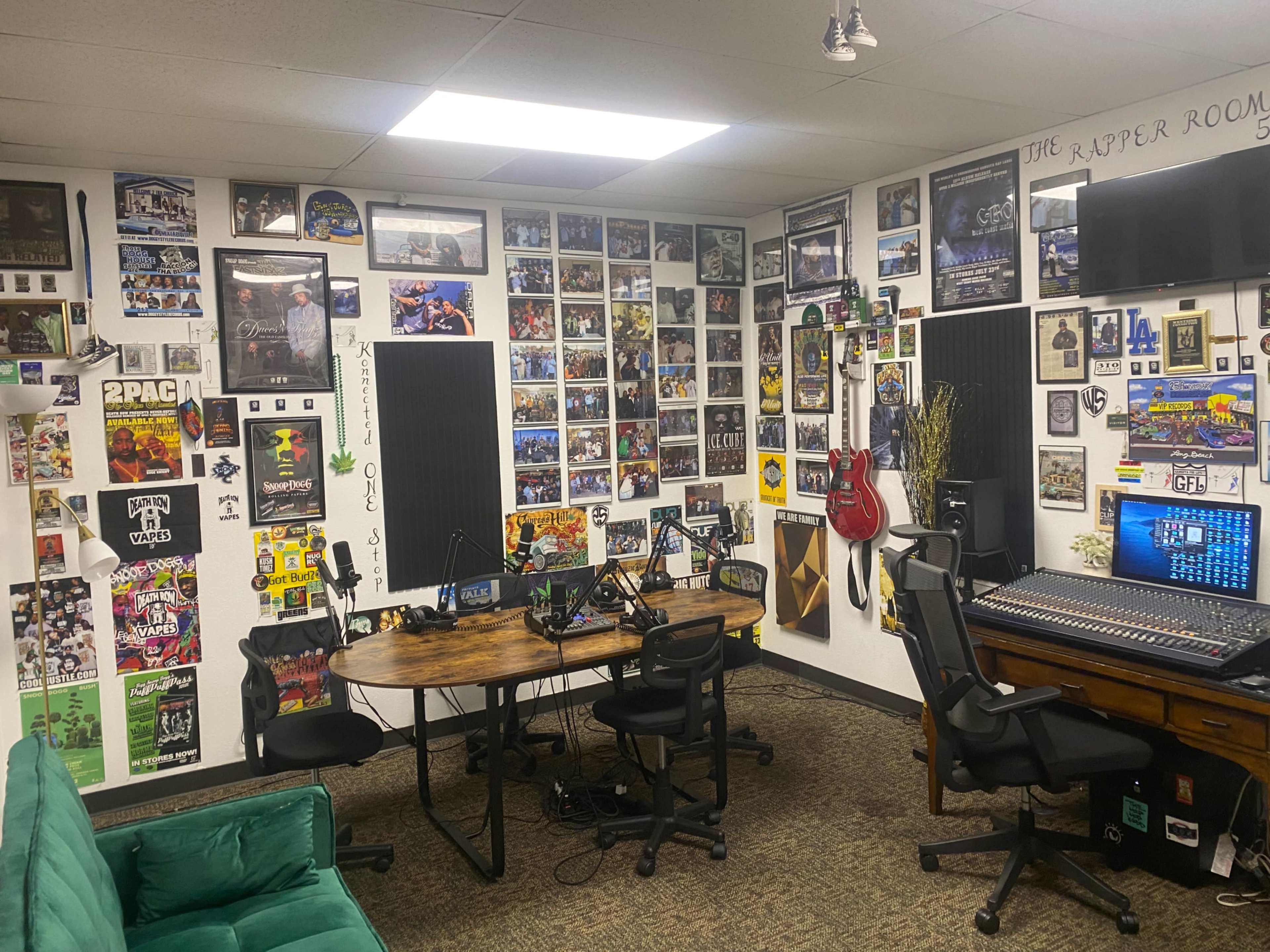 The image shows a recording studio with a circular wooden table and chairs, surrounded by walls decorated with numerous framed photos and posters, and a sound mixing console on one side.