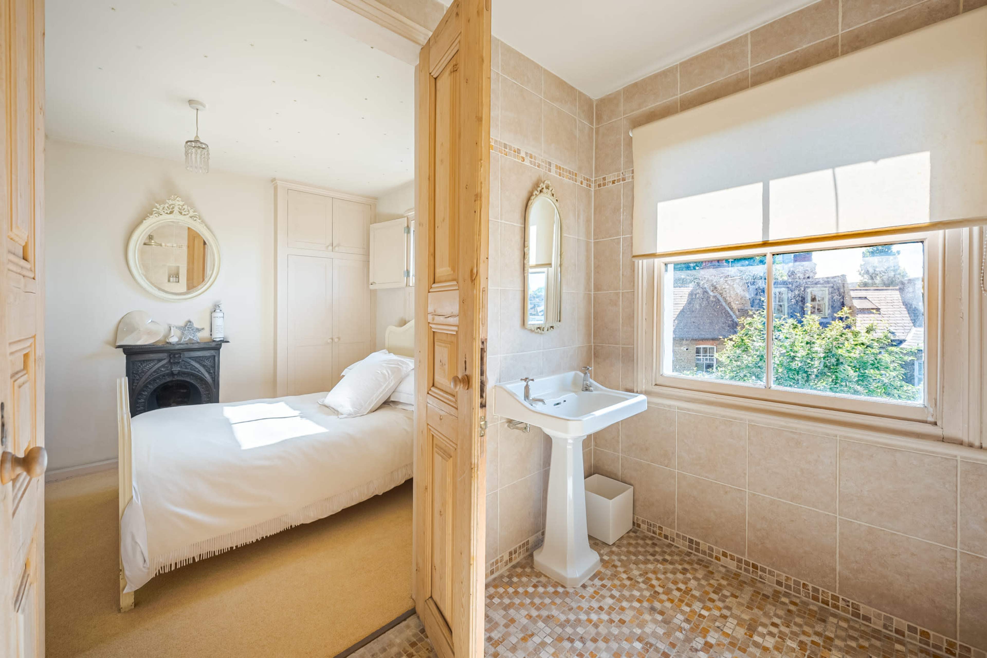 The image shows a bright bedroom with a wooden door leading to a bathroom featuring a sink and a window overlooking a tree-lined view.