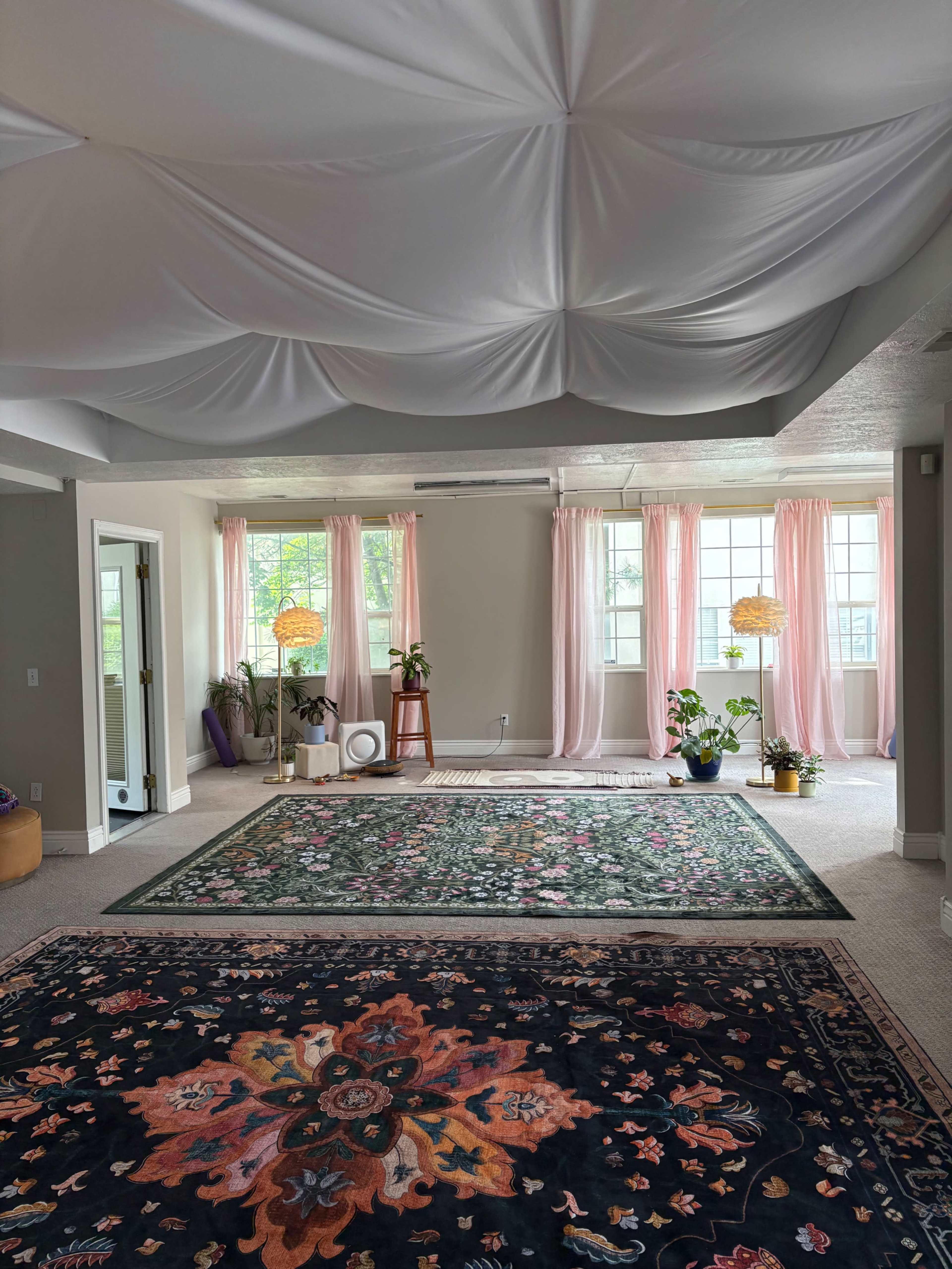 The image shows a spacious room with draped white fabric ceiling, pastel curtains, plants in various pots, and two large decorative rugs on the floor.