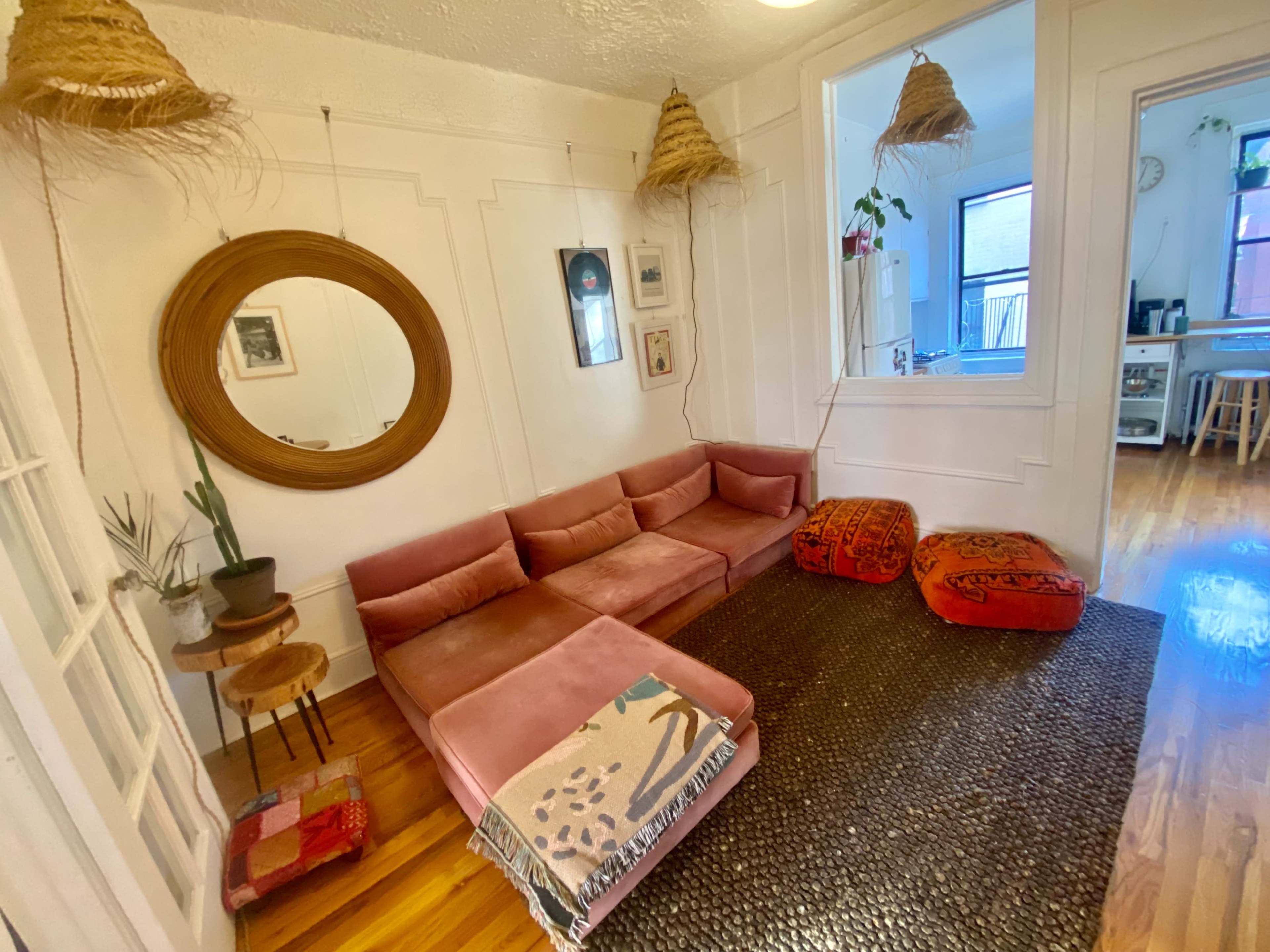 A living room features a pink sectional sofa, a round mirror, a woven rug, and decorative poufs, with plants and wall art in a well-lit space.