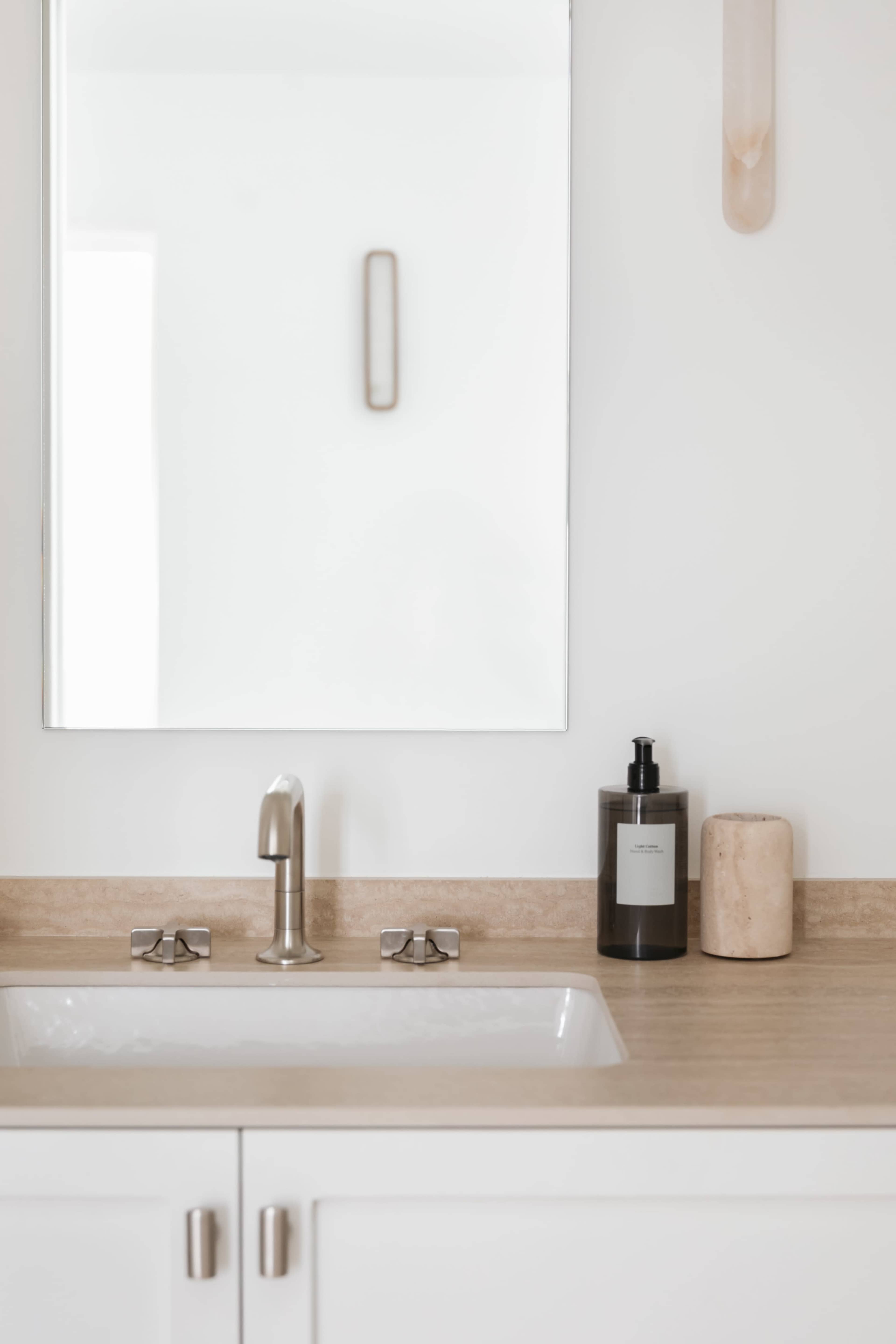 A bathroom counter features a sink, a mirror, a soap dispenser, and a stone container against a light-colored wall.