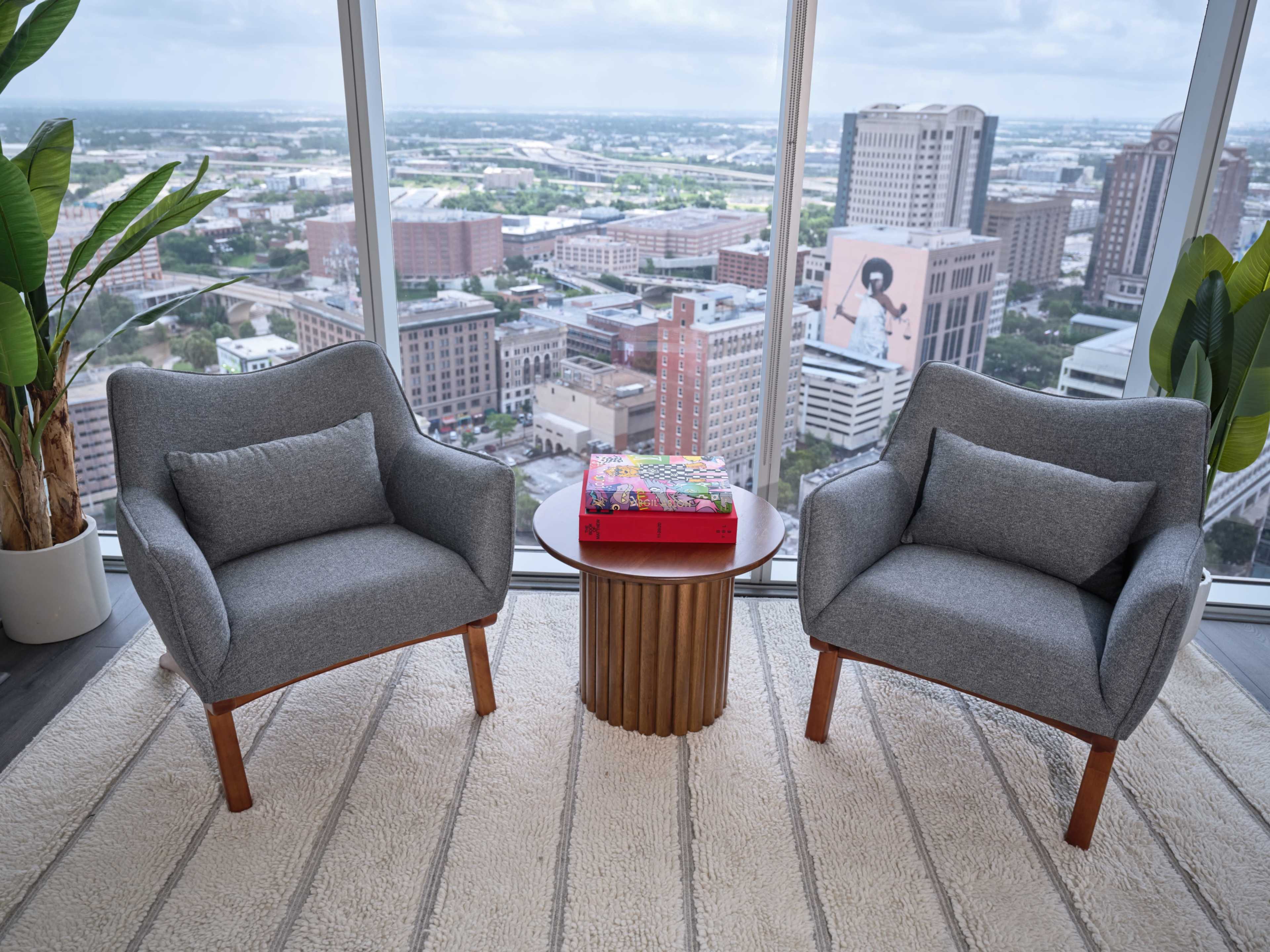 Two gray armchairs are positioned on a rug across from a round wooden coffee table, overlooking a city skyline through large windows.