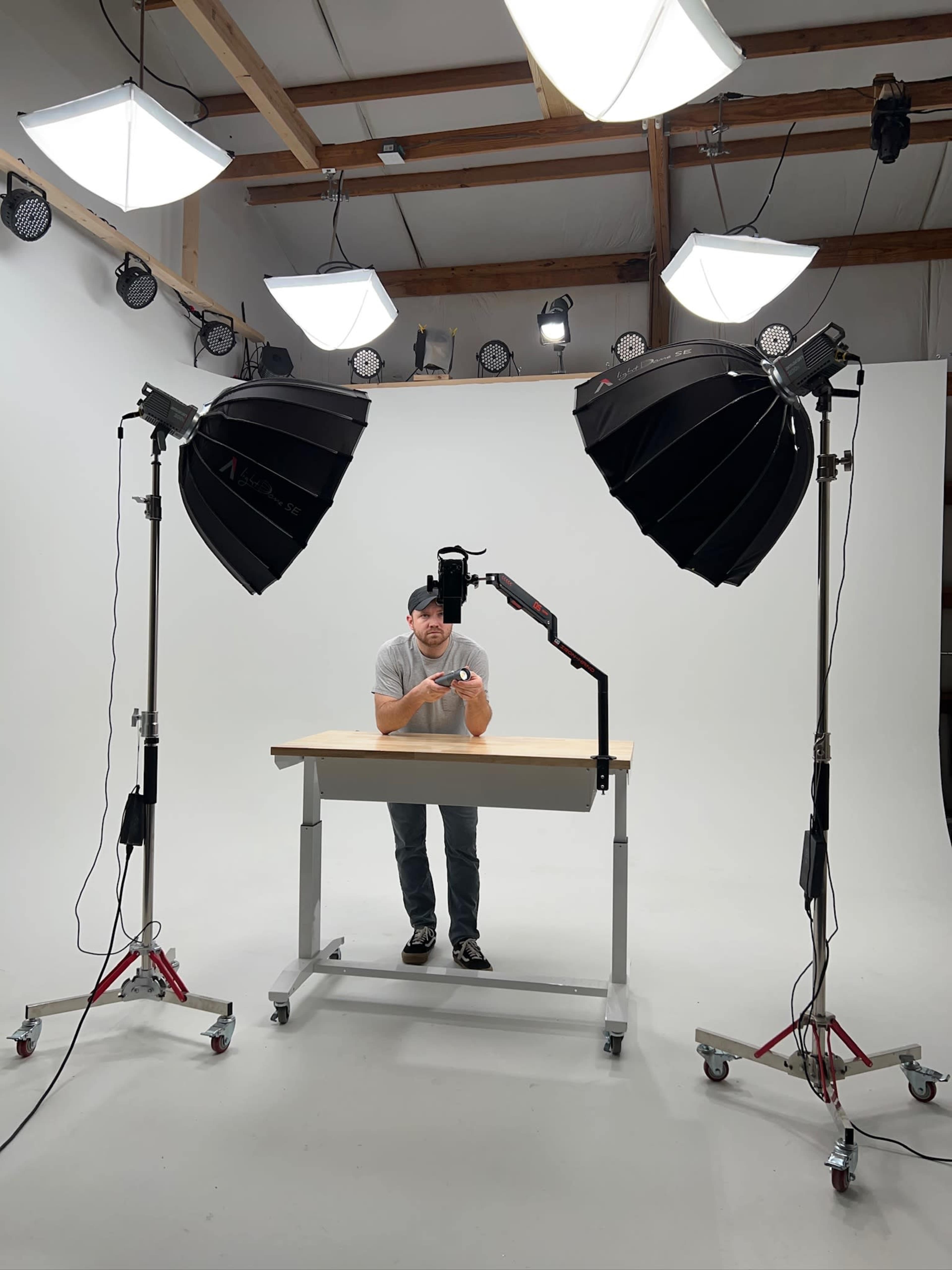 A person stands at a table in a well-lit photography studio, surrounded by large softbox lights and a blank white backdrop.