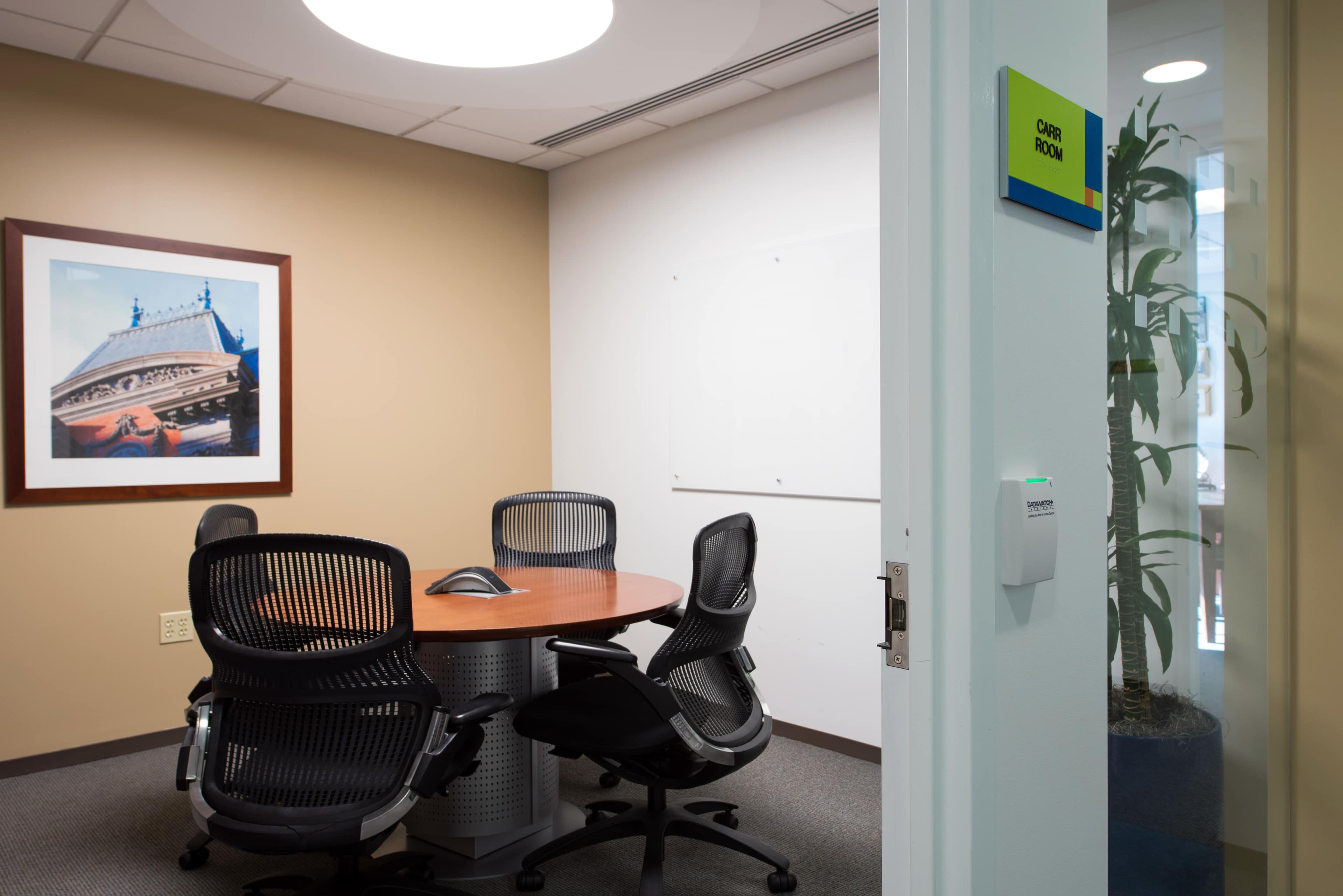 A small conference room features a round wooden table surrounded by black ergonomic chairs, with a large framed photograph on the wall and a glass door showing a potted plant.