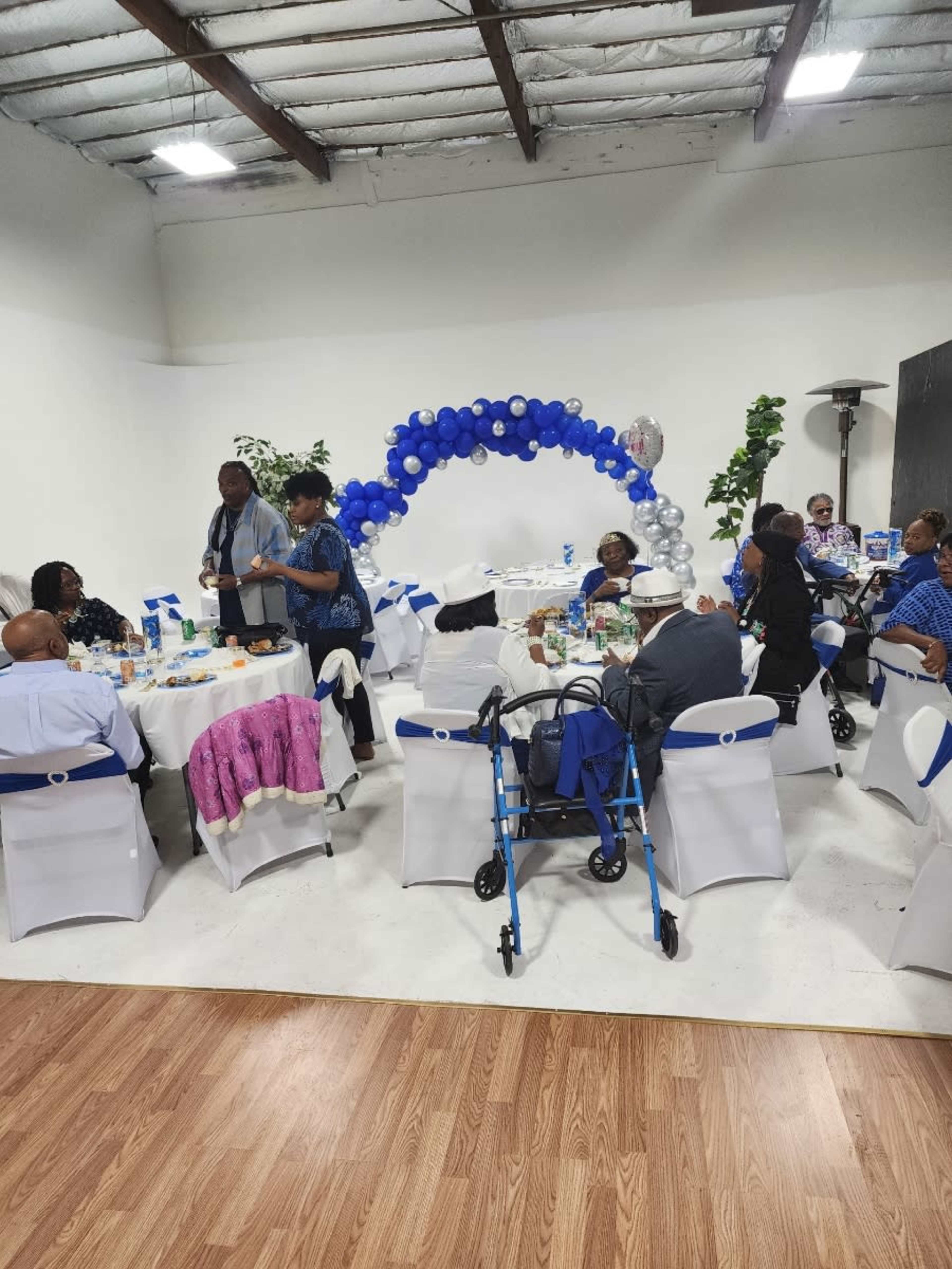 A group of people is gathered in a decorated room with tables set for an event, featuring blue and silver balloons in an archway.