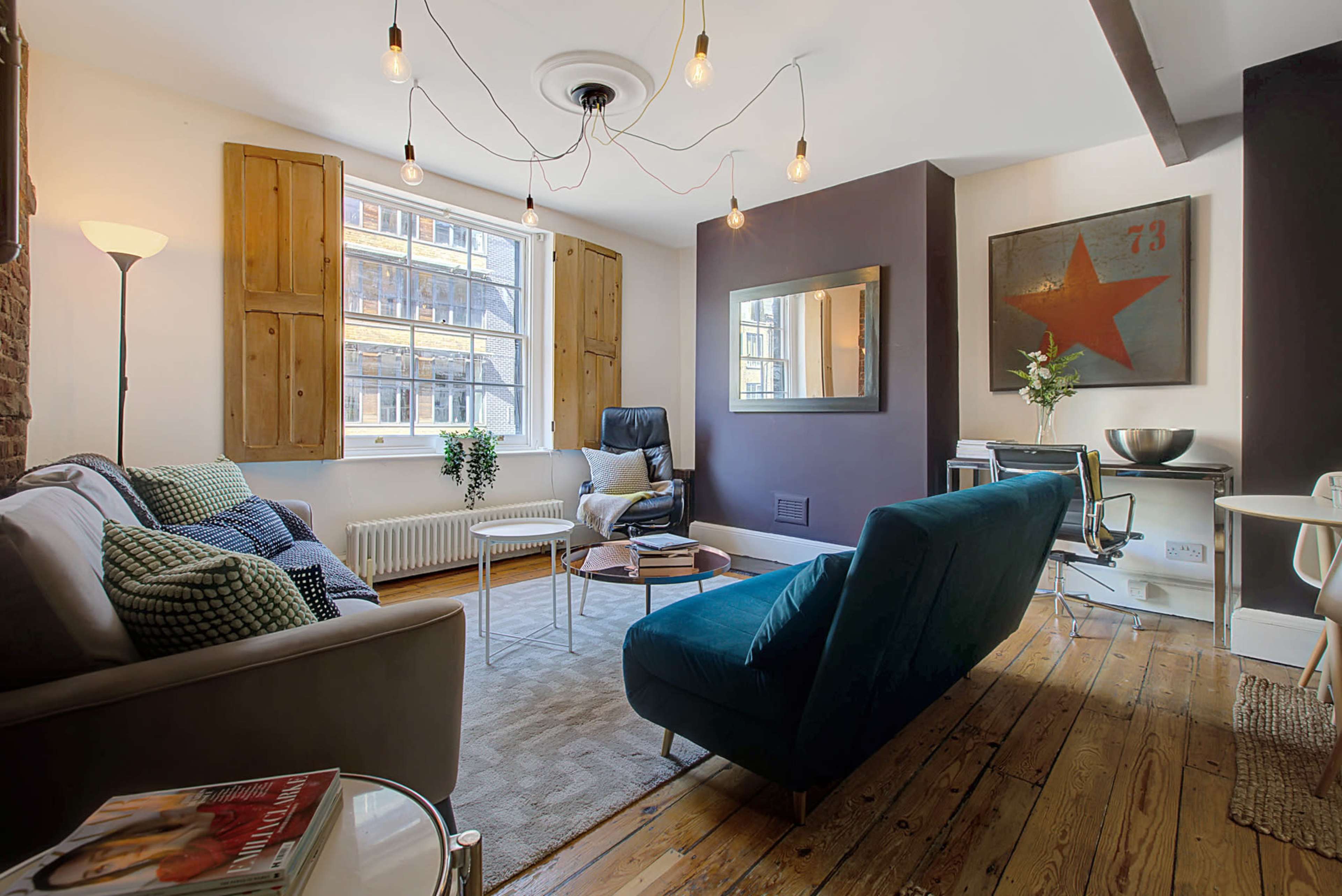 The image shows a modern living room with wooden floors and a mix of furniture, including a sofa, armchair, and a desk,along with large windows letting in natural light.