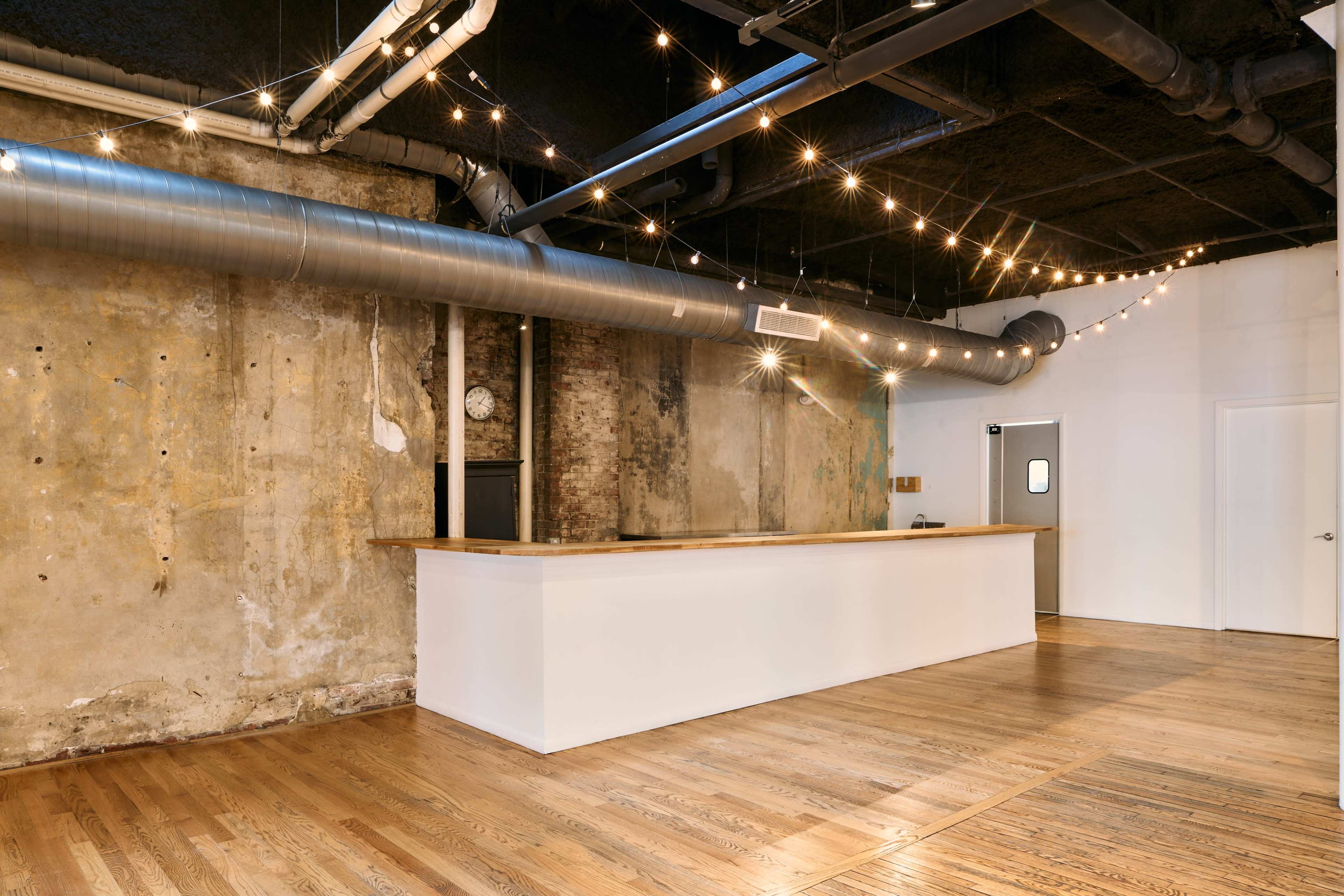 The image shows a spacious room with a wooden counter against a textured wall, overhead string lights, and exposed pipes.
