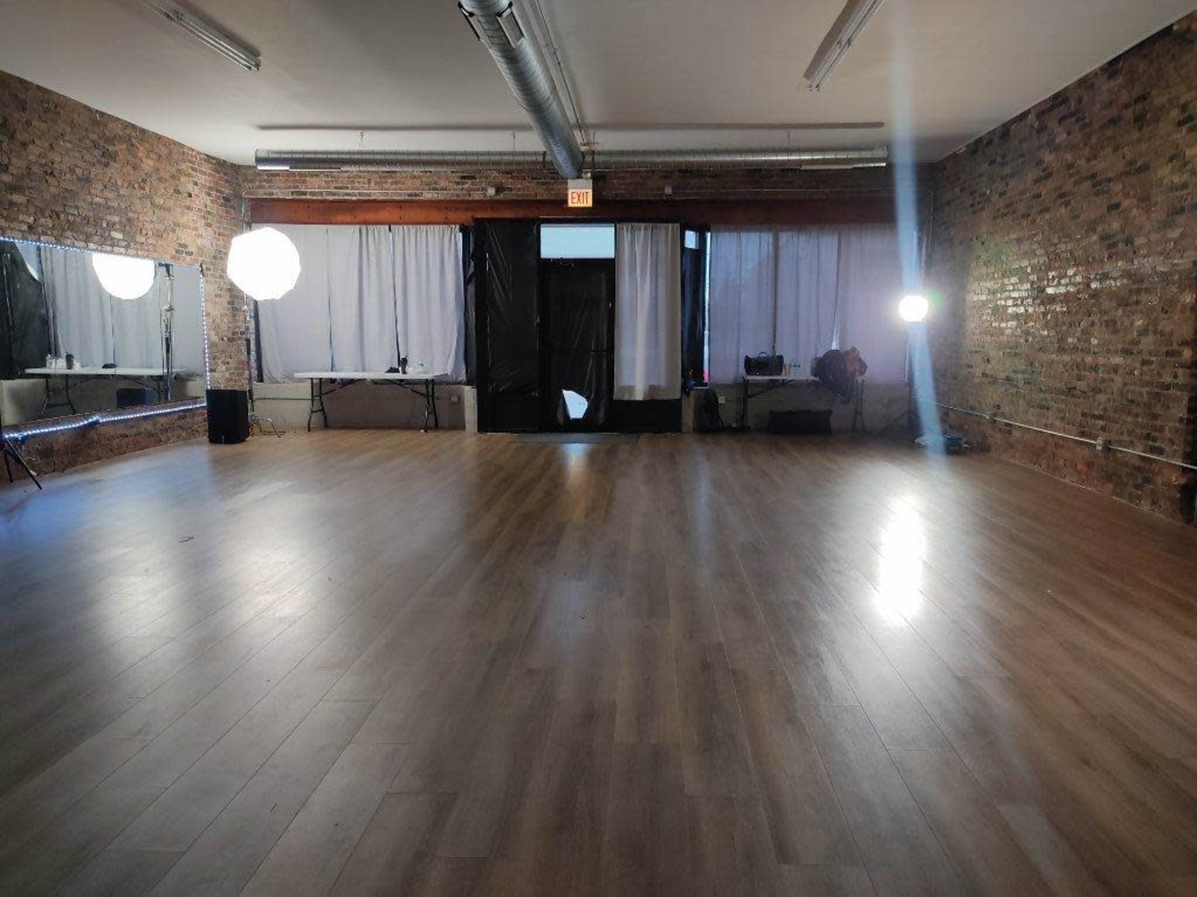 A spacious empty studio with wooden flooring, exposed brick walls, and large windows covered by white curtains, illuminated by two softbox lights.