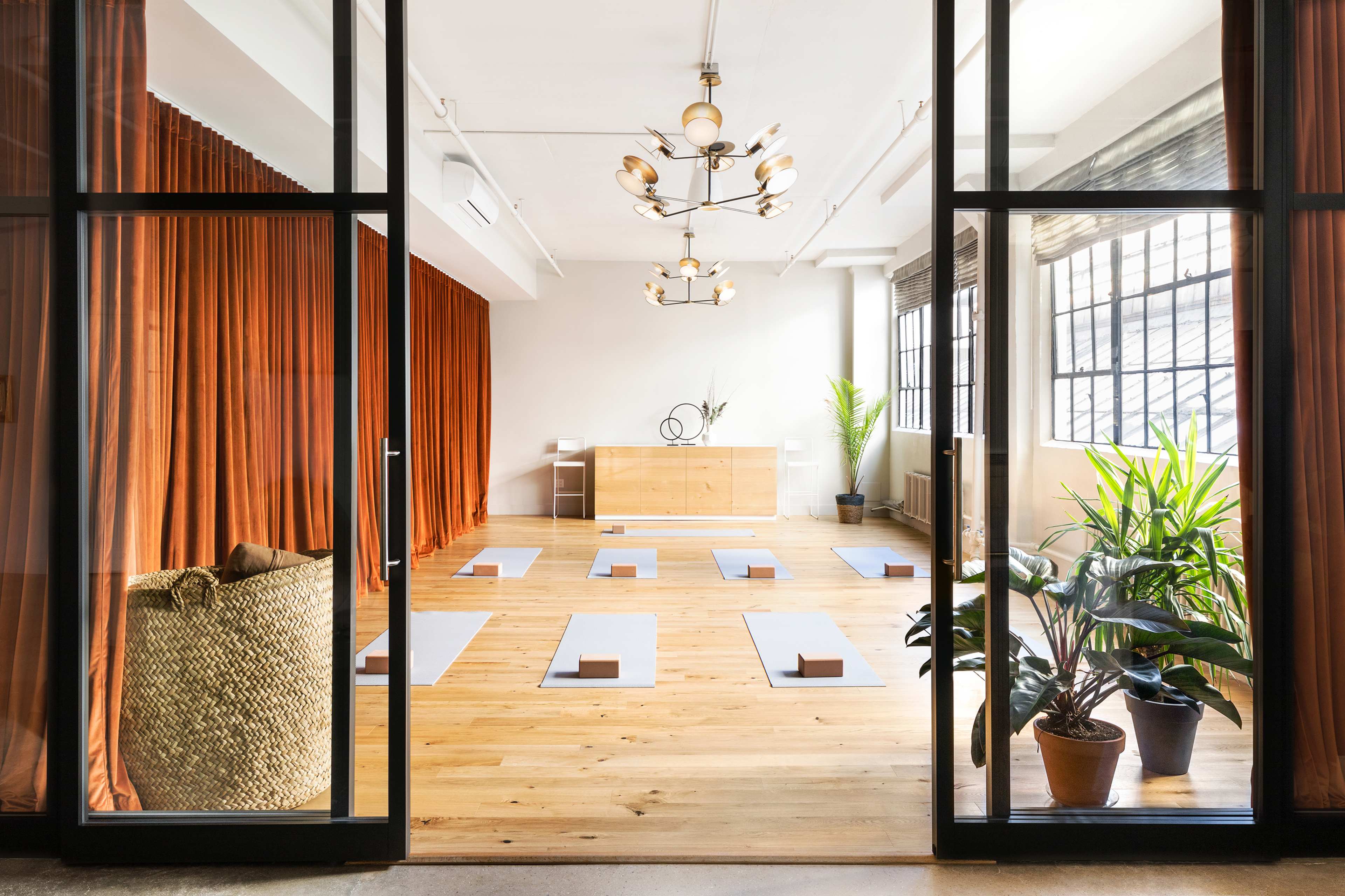 A bright yoga studio features wooden flooring, large windows, and several mats arranged neatly on the floor, surrounded by green plants and soft drapery.