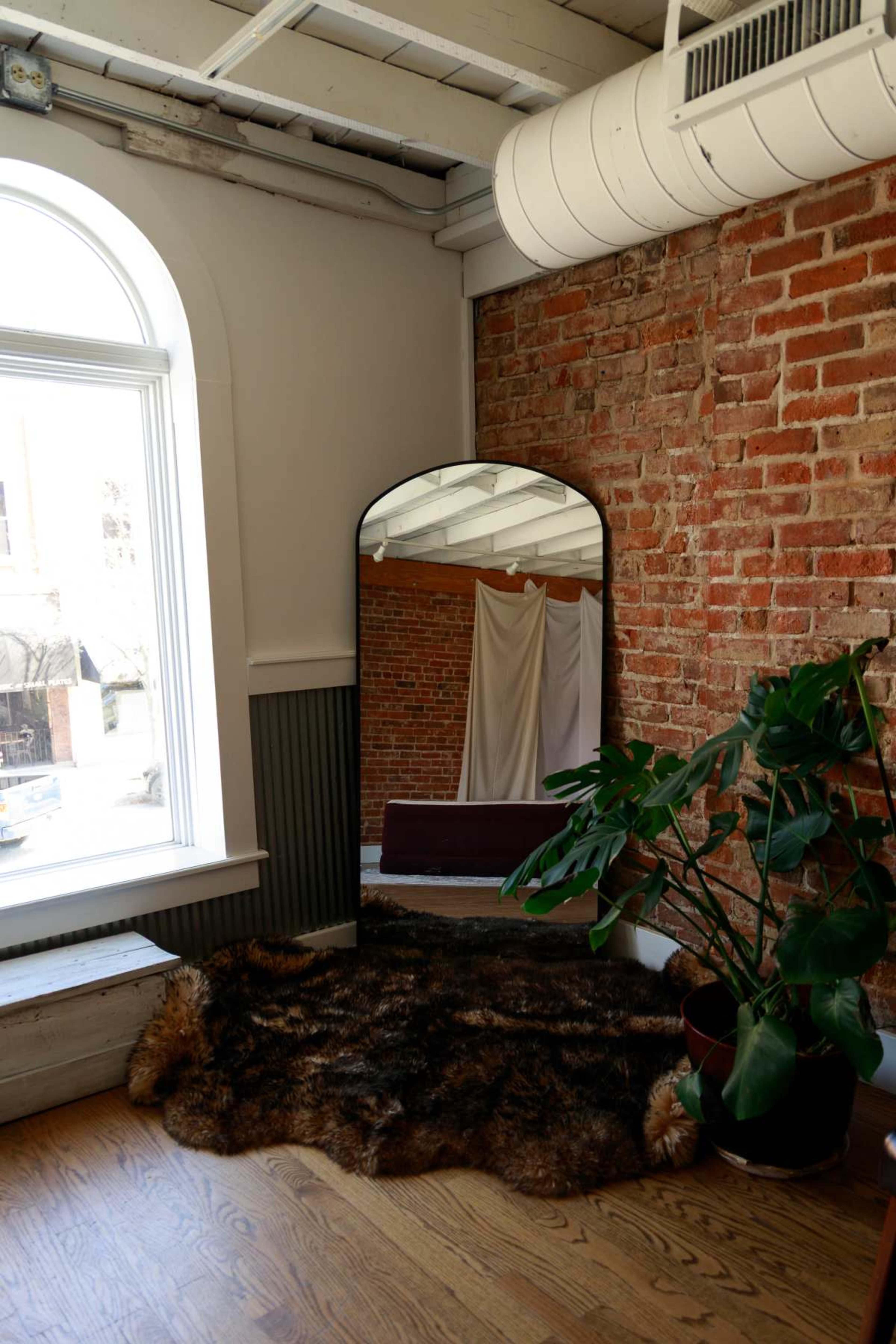 The image features a large arched mirror leaned against a brick wall in a room with wooden flooring and a potted plant on a fur rug.