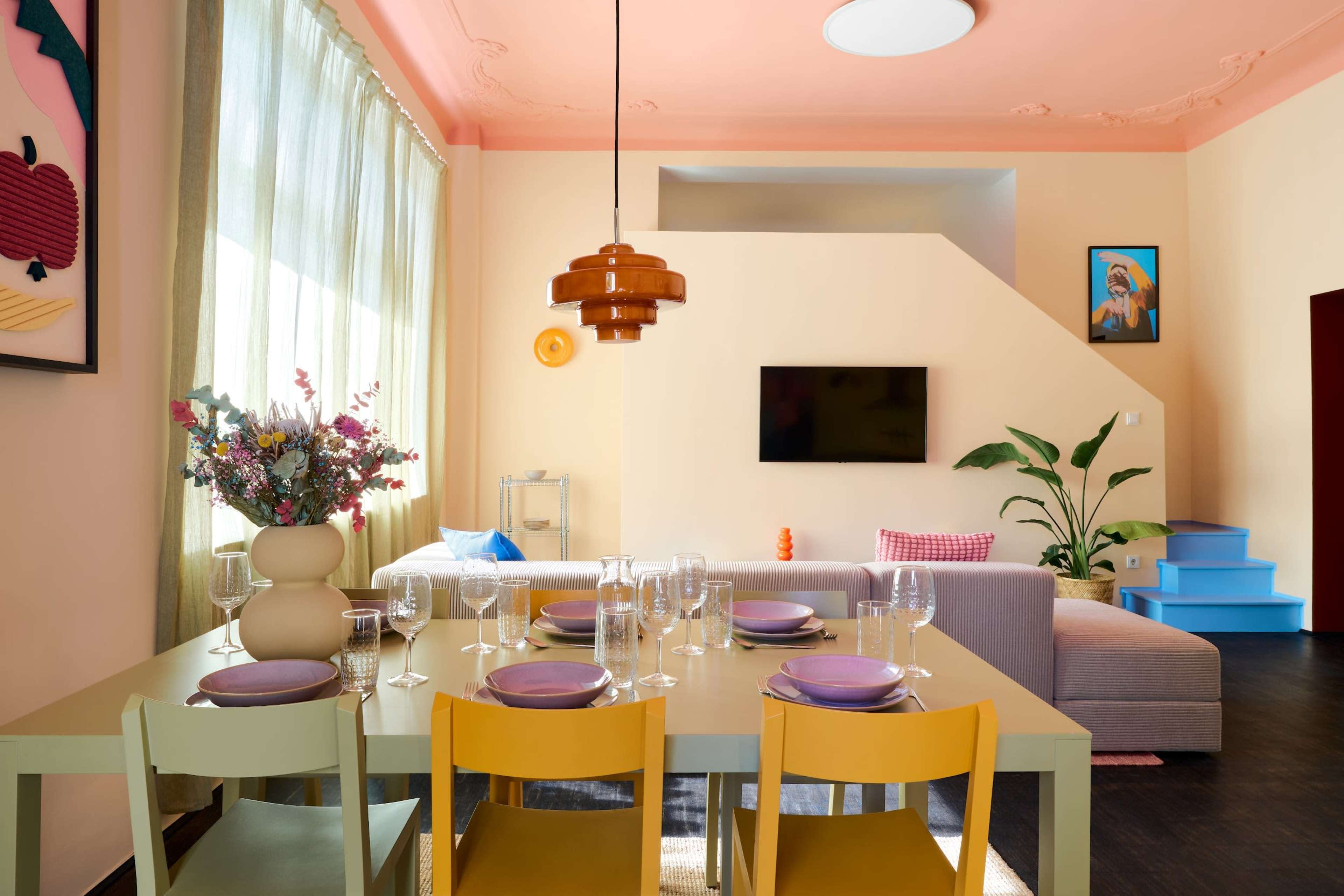 A dining area features a multicolored table set with plates and glasses, surrounded by a couch and a modern plant, under soft natural light.