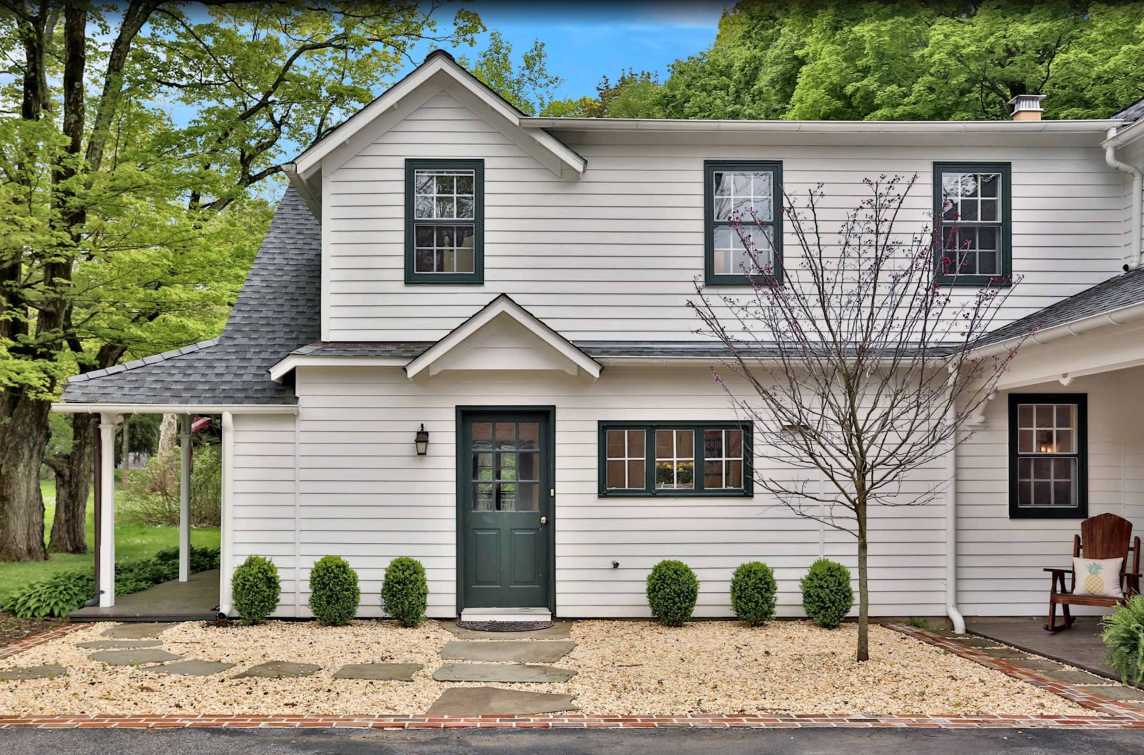 A two-story white house with dark green accents features a stone pathway leading to the front door, surrounded by neatly trimmed shrubs and gravel.