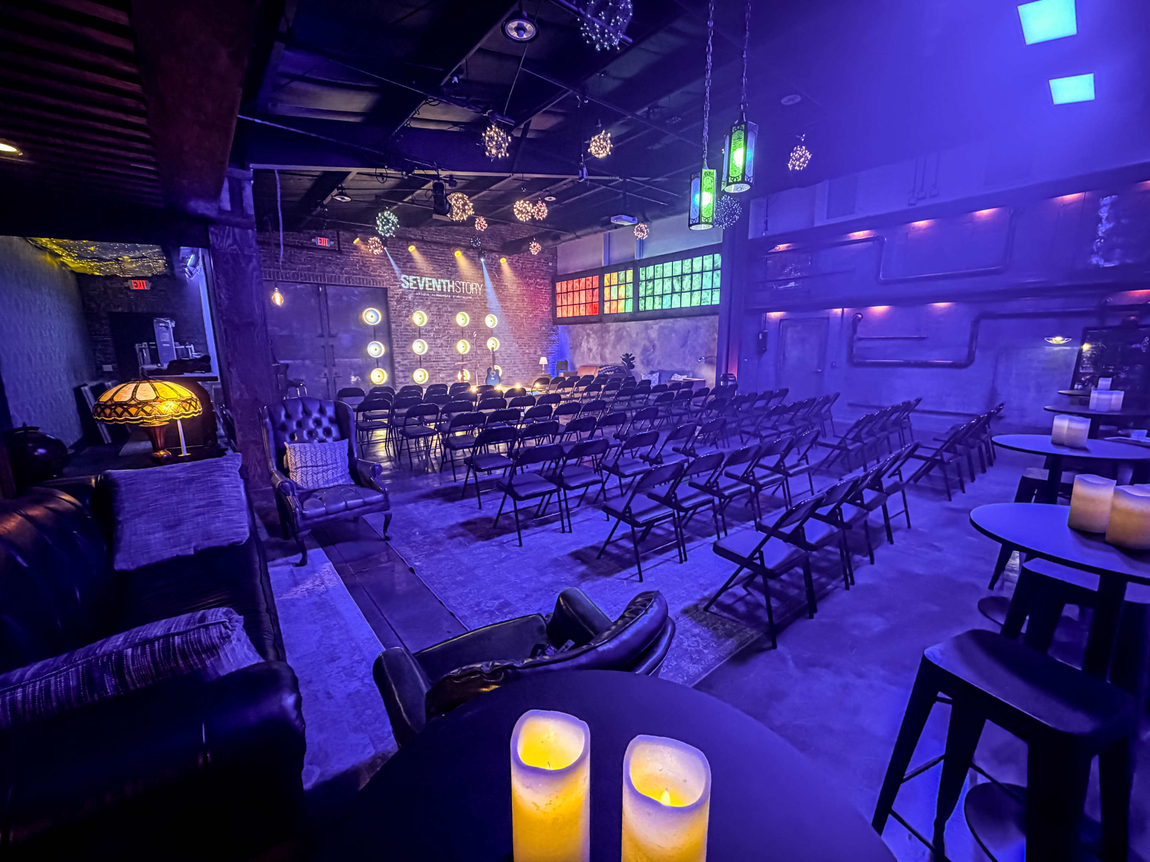 The interior of a dimly lit venue features rows of empty chairs facing a stage, with decorative lighting and candles on tables.