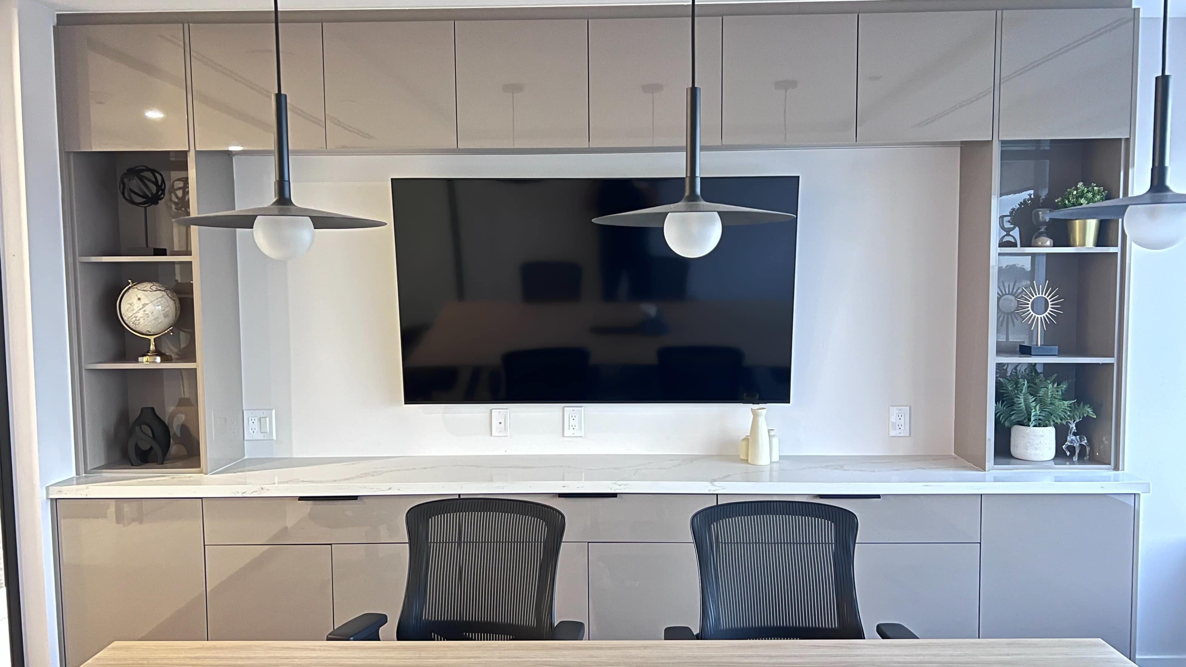 The image shows a modern office space featuring a wall-mounted television, two black chairs, and shelving with decorative items and lighting fixtures.