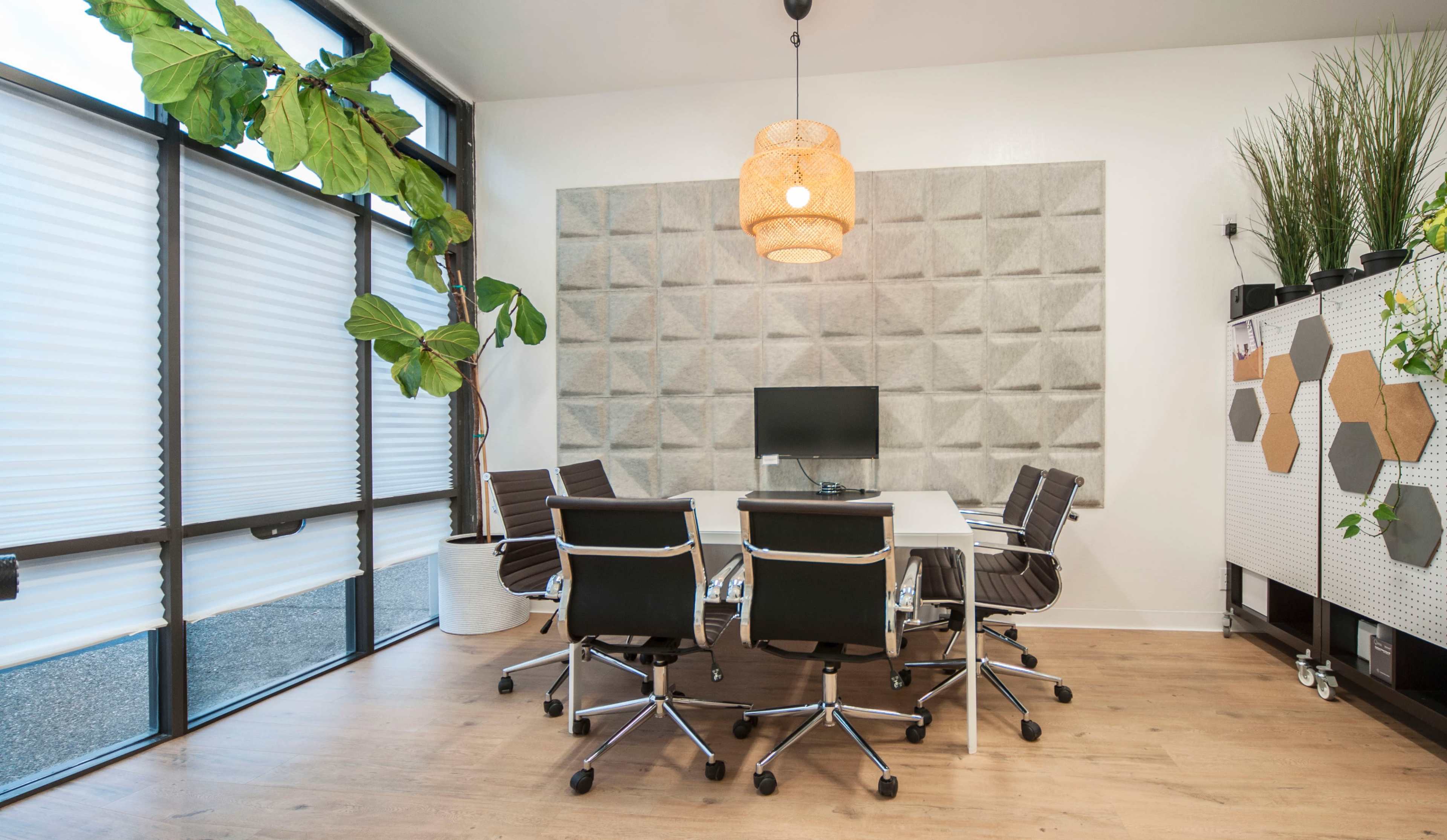 A modern conference room features a round white table surrounded by black swivel chairs, with a wall-mounted monitor and a large plant near a textured gray wall.