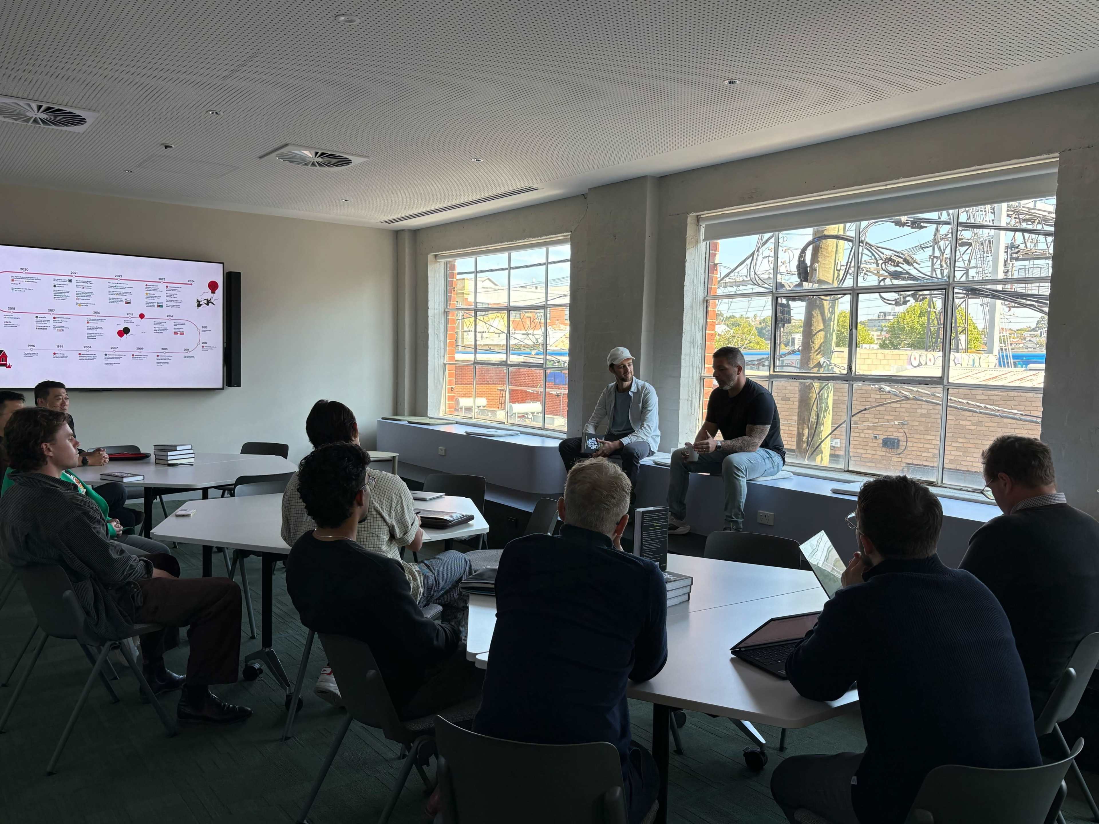 Two speakers are seated in front of a group of attendees, who are participating in a discussion in a brightly lit room with large windows.