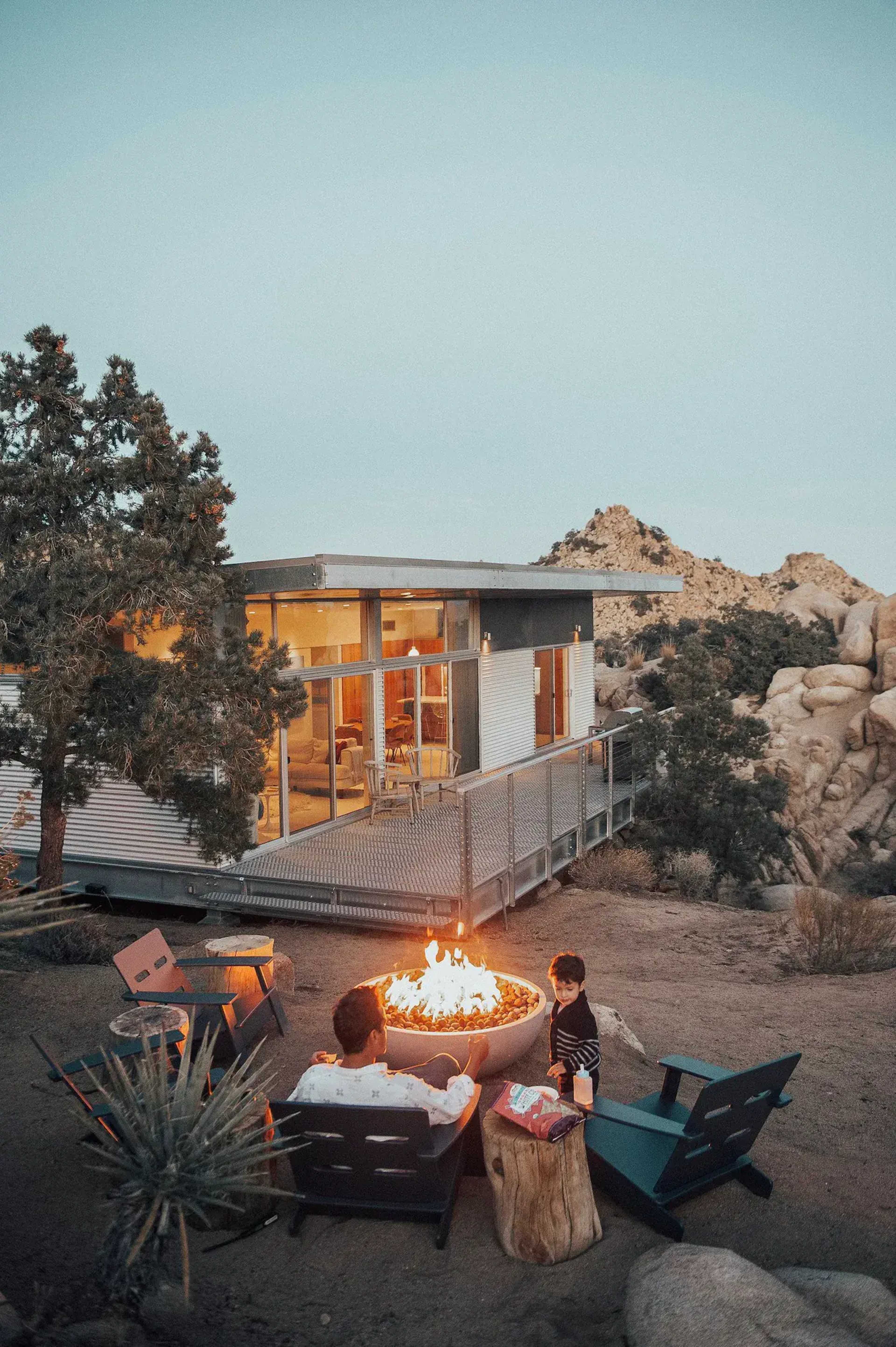 A man and a child sit around a fire pit in a desert landscape, with a modern glass house and rocky terrain in the background.