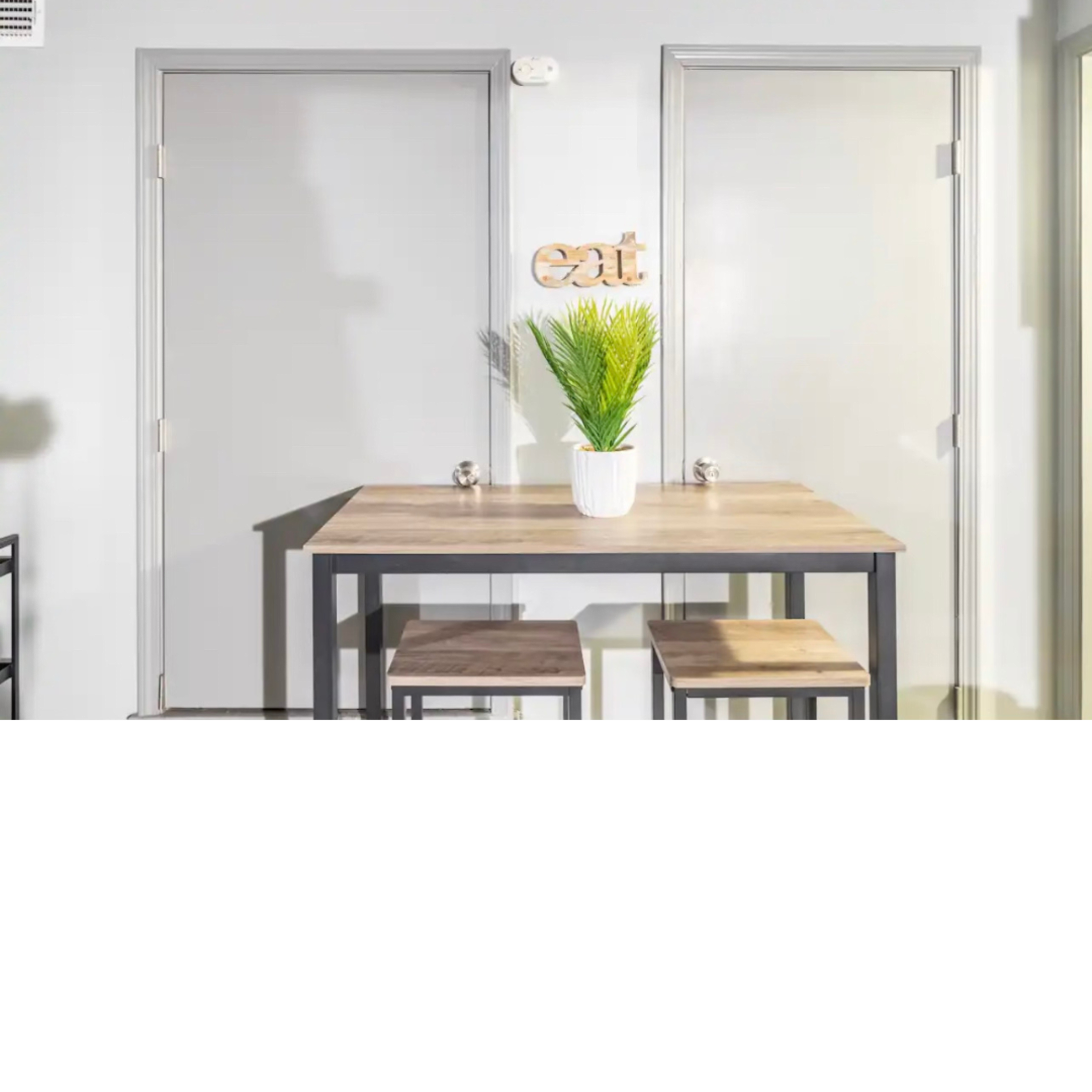 A small dining area features a wooden table with two stools and a potted plant, flanked by two closed doors.
