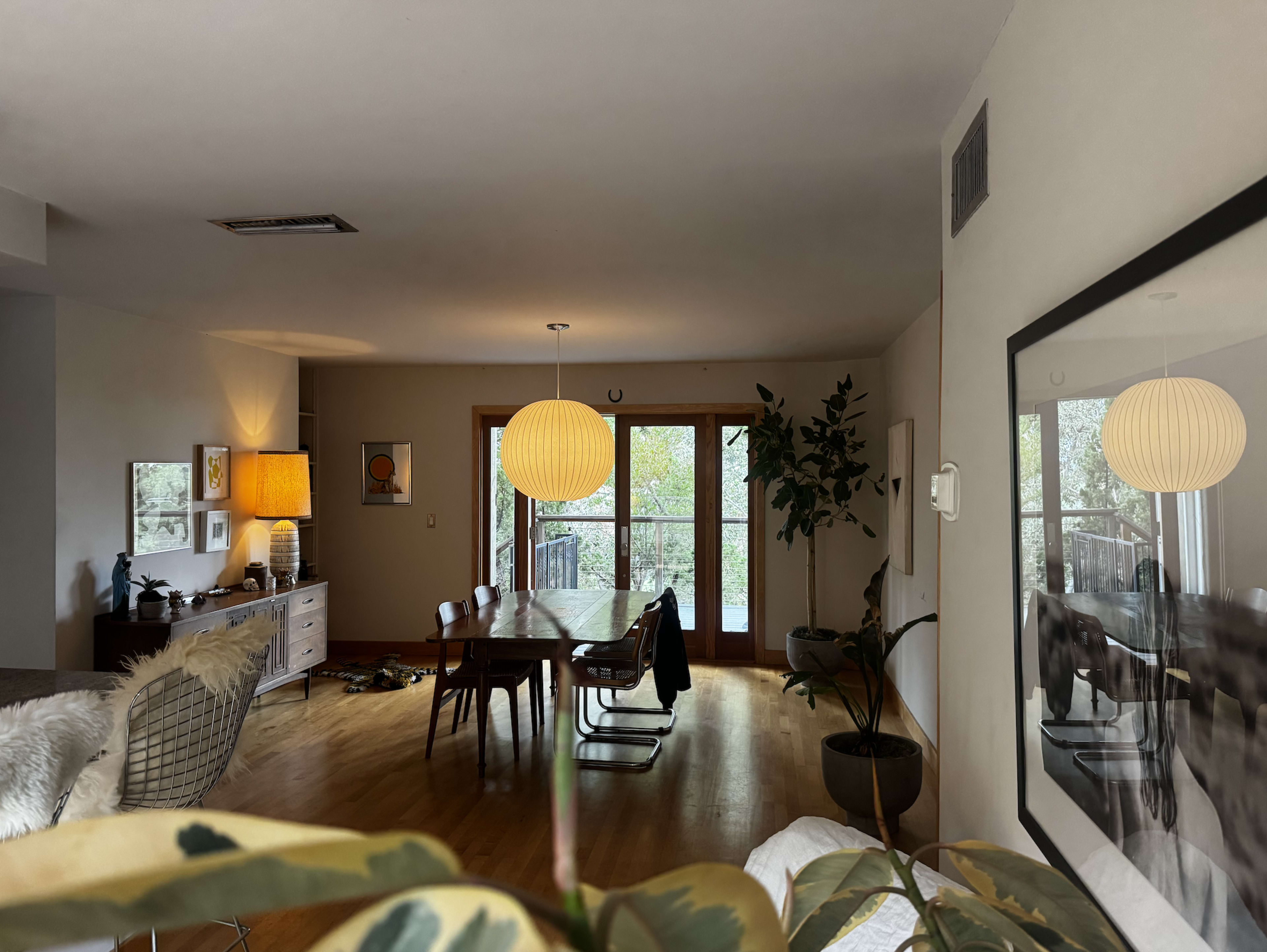 The image shows a modern living space with a dining area, large windows, plants, and a light fixture hanging from the ceiling.