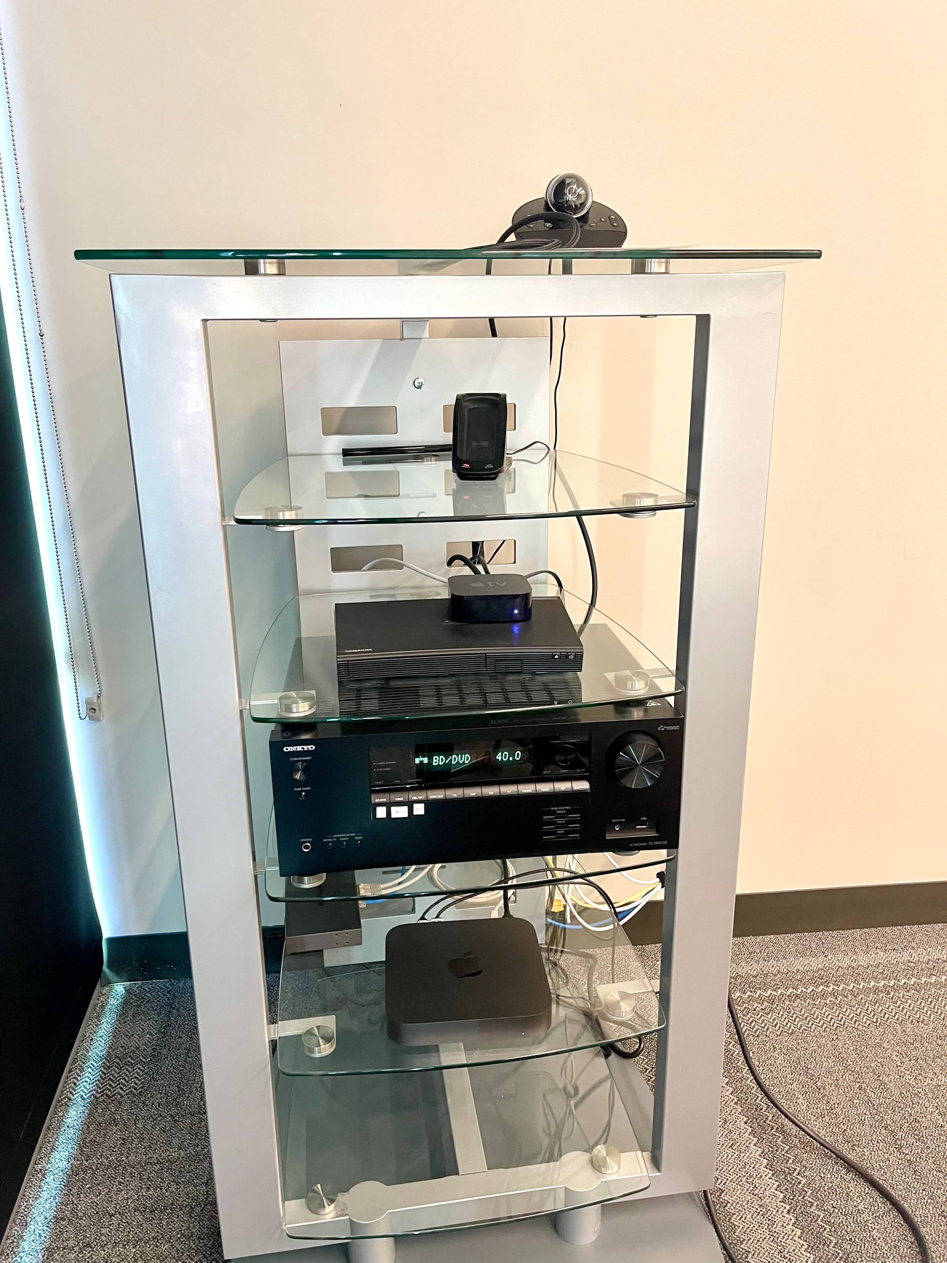 The image shows a modern audio-visual equipment rack with a glass top, containing various electronic devices, including a webcam, a DVD player, and a media streaming device.