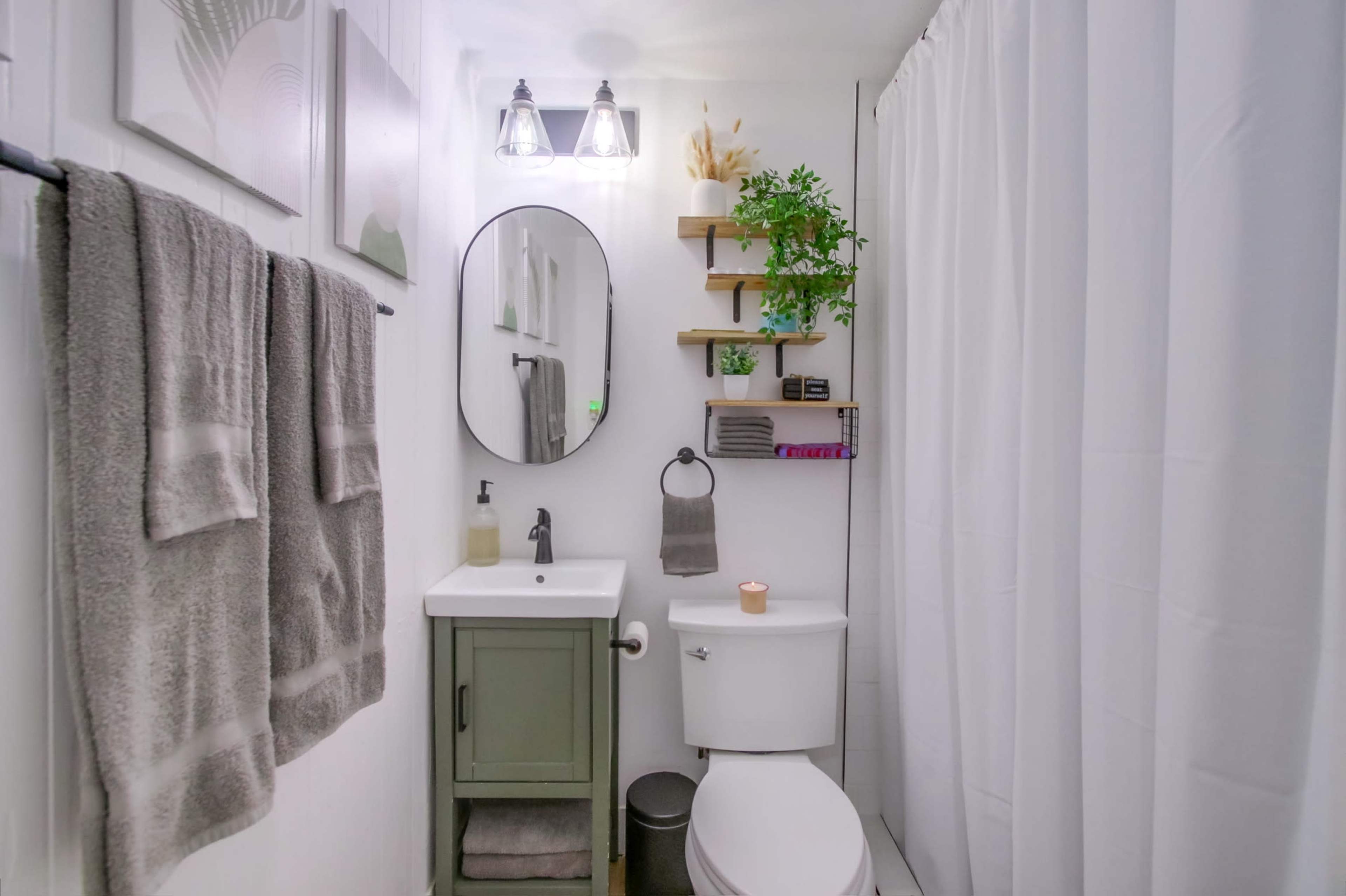 The image shows a compact bathroom featuring a mirror above a small sink vanity, towels hanging on the wall, and a shower curtain made of white fabric.