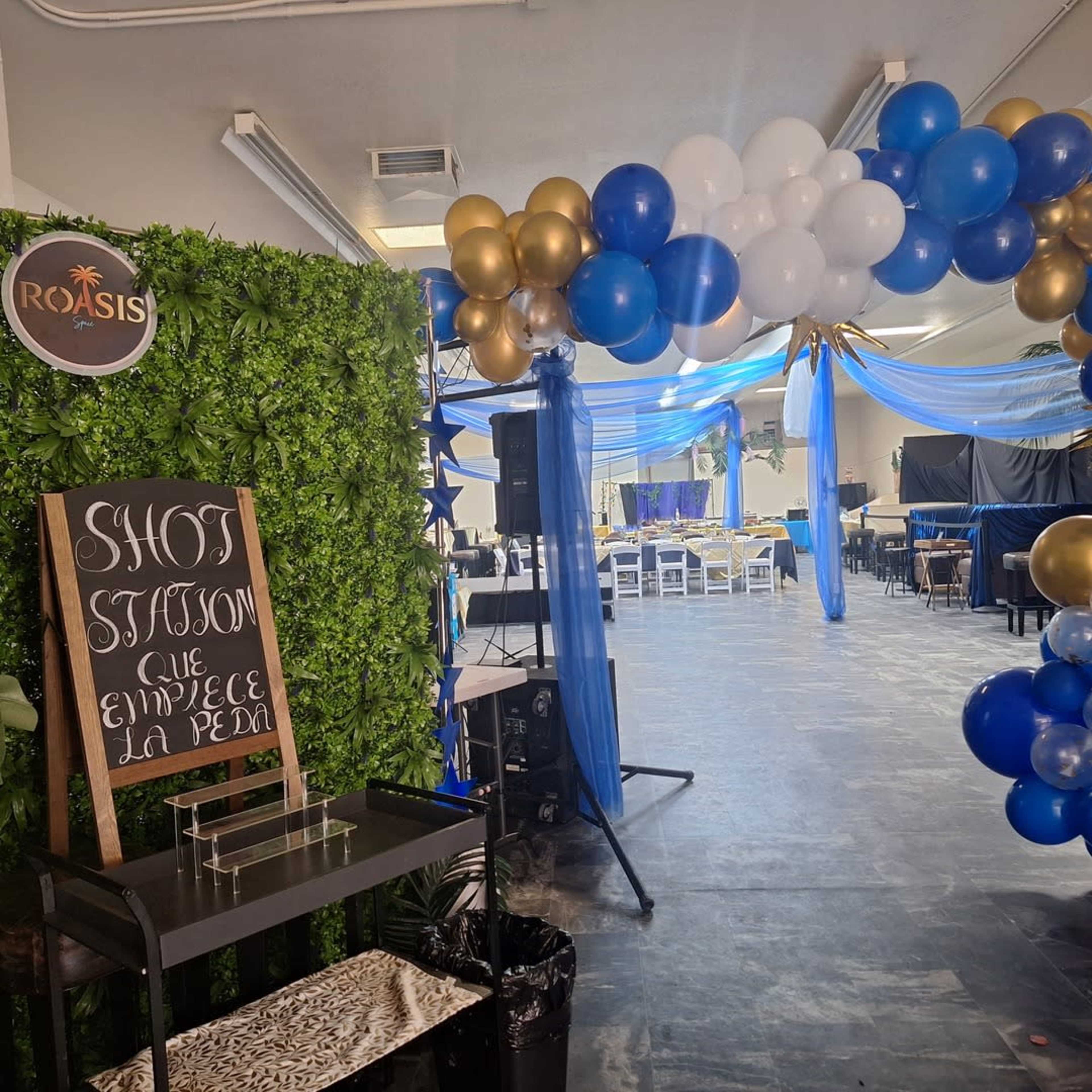 The image shows a decorated event space with a green wall, a sign for a shots station, and a balloon arch leading to a gathering area set up for a celebration.