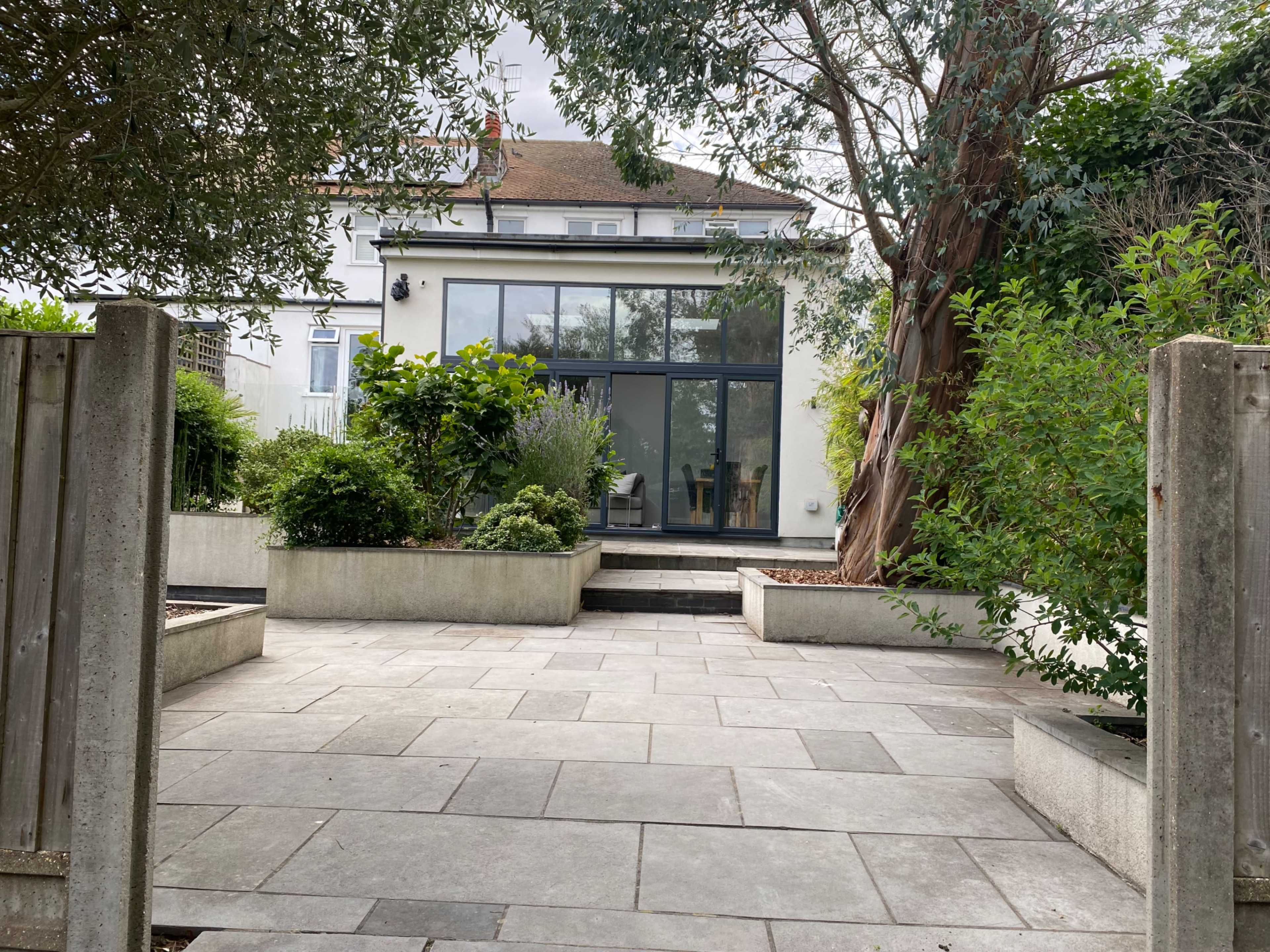 The image shows a modern home with large windows surrounded by landscaped greenery and a stone-paved patio in the foreground.