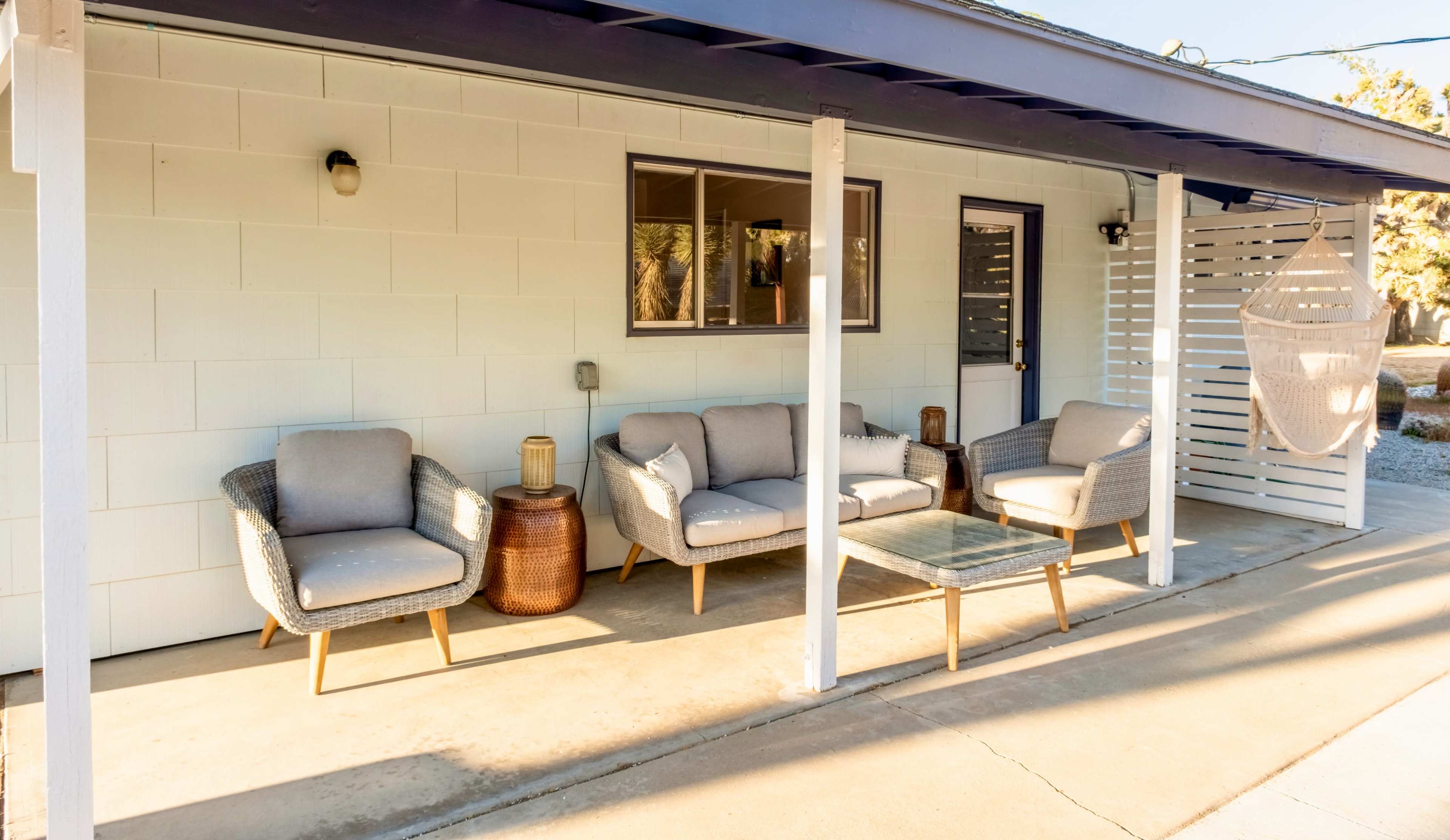 A covered outdoor seating area with a glass table, two armchairs, and a couch, situated beside a wall.