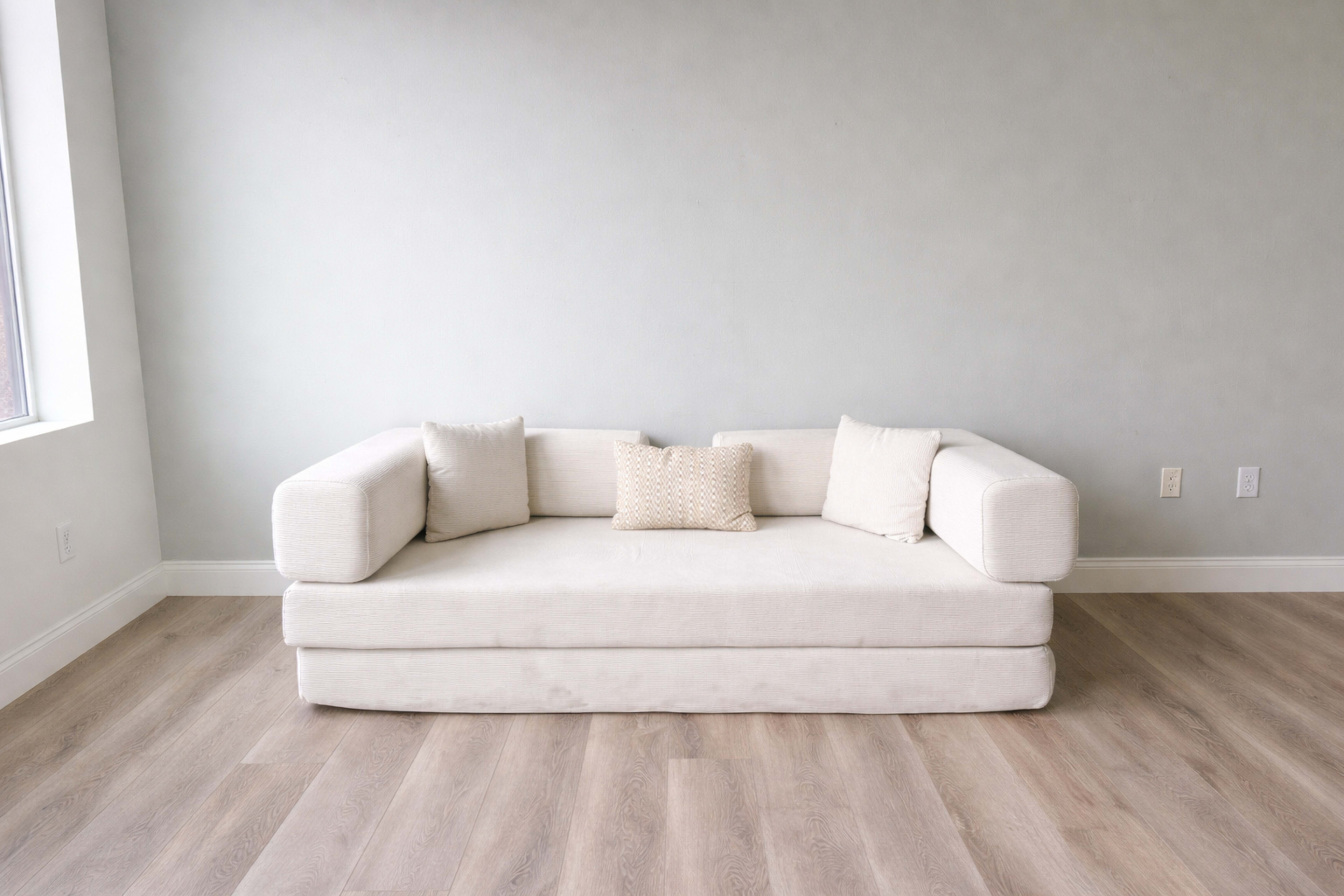 A light-colored sofa with two armrests and three cushions is positioned against a gray wall on a wooden floor.