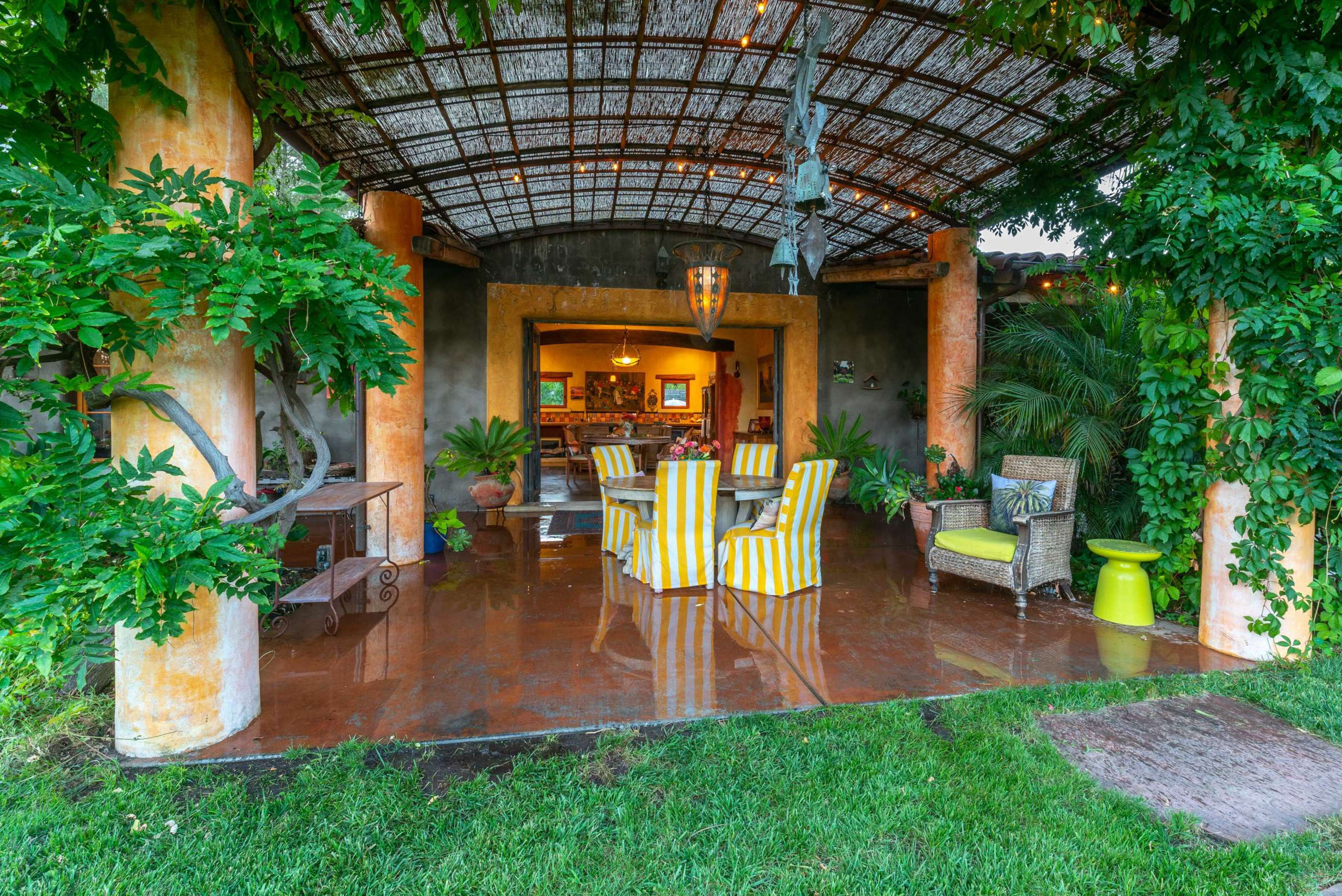 A covered patio area with striped chairs and a wooden table, surrounded by greenery and decorative plants.