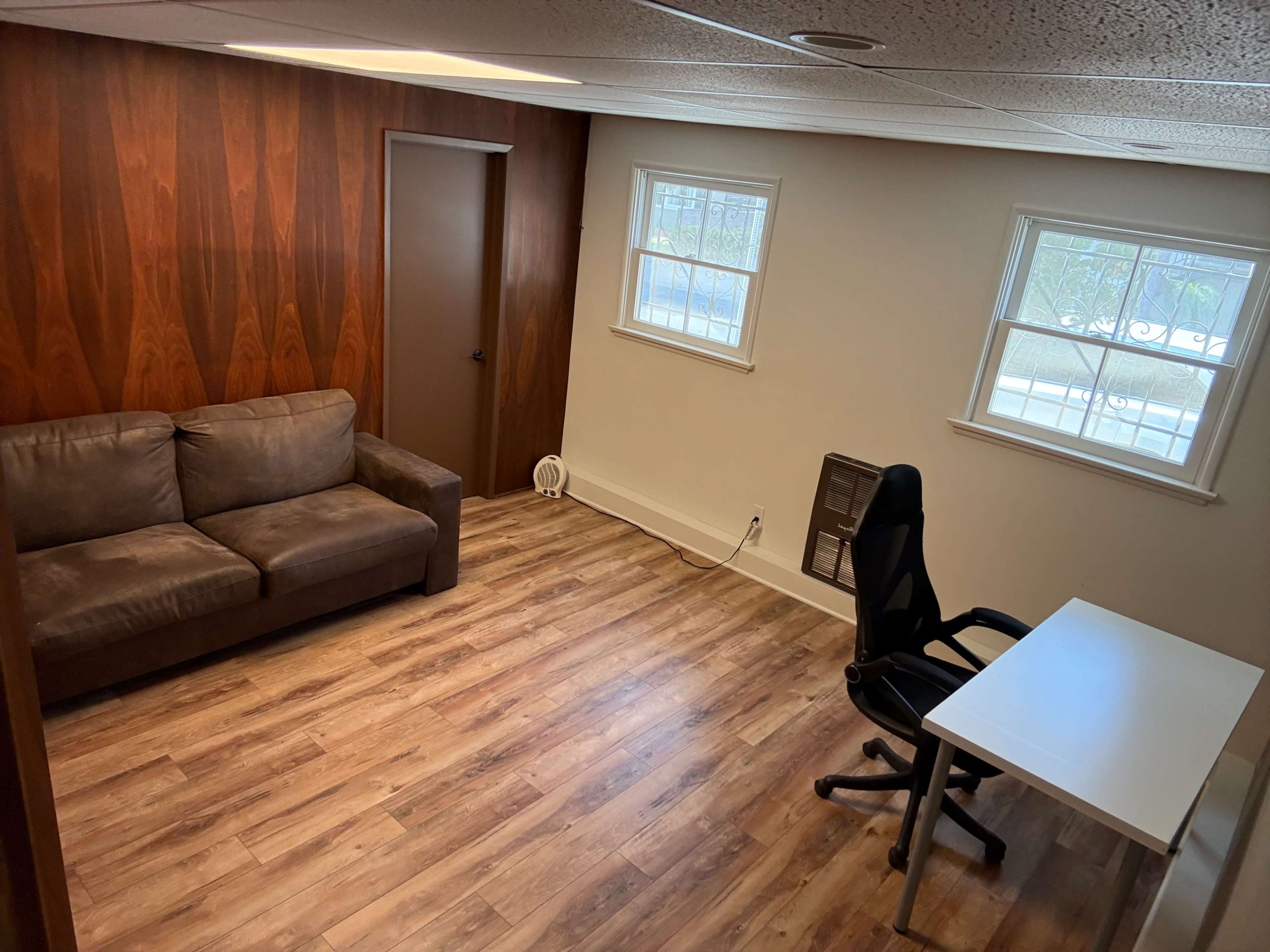 The image shows a room with a brown sofa, a black office chair, and a white desk, surrounded by wooden paneling and two windows.