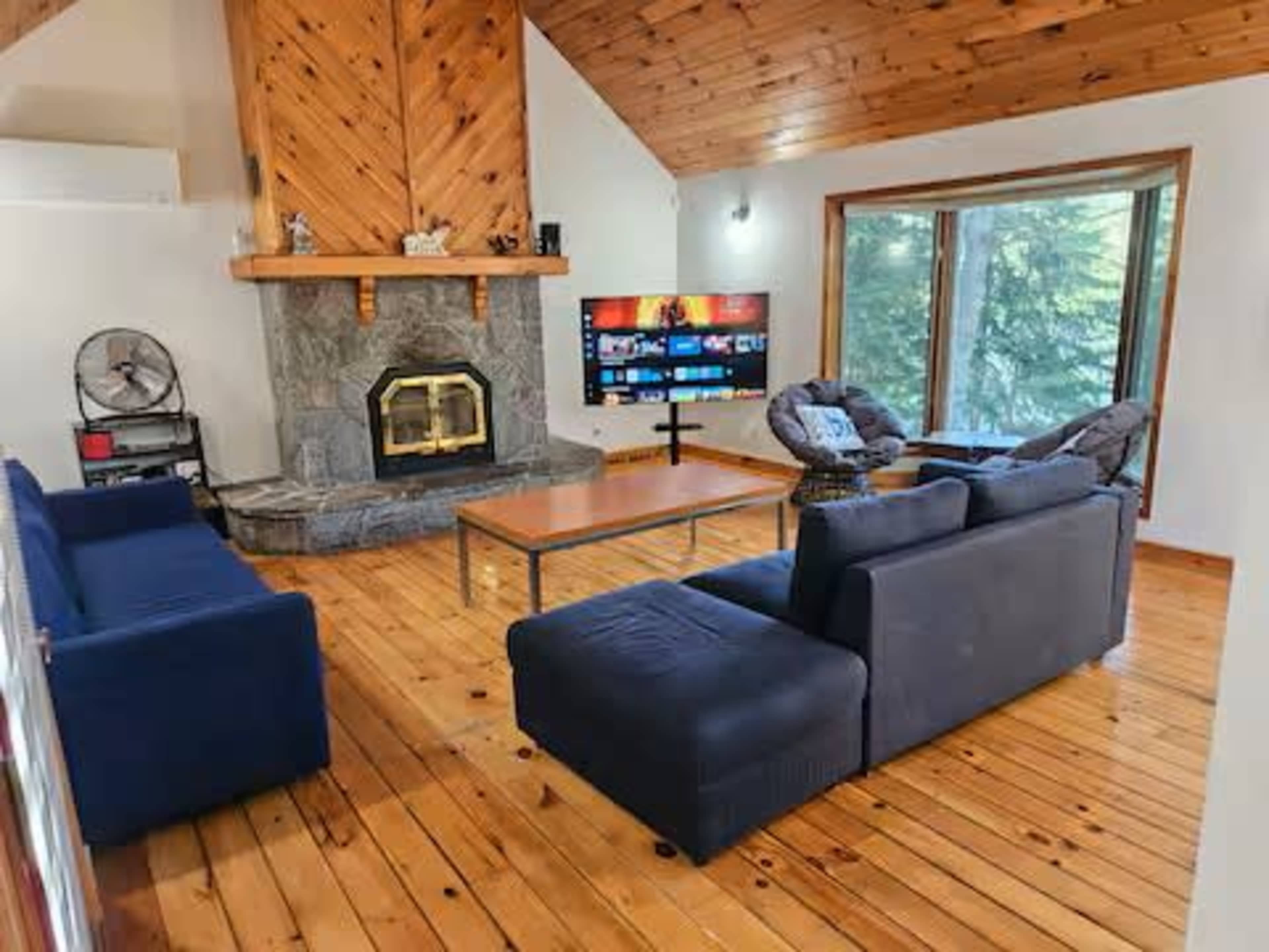 The image shows a cozy living room with a stone fireplace, wooden ceiling, and seating arranged around a coffee table and a television.