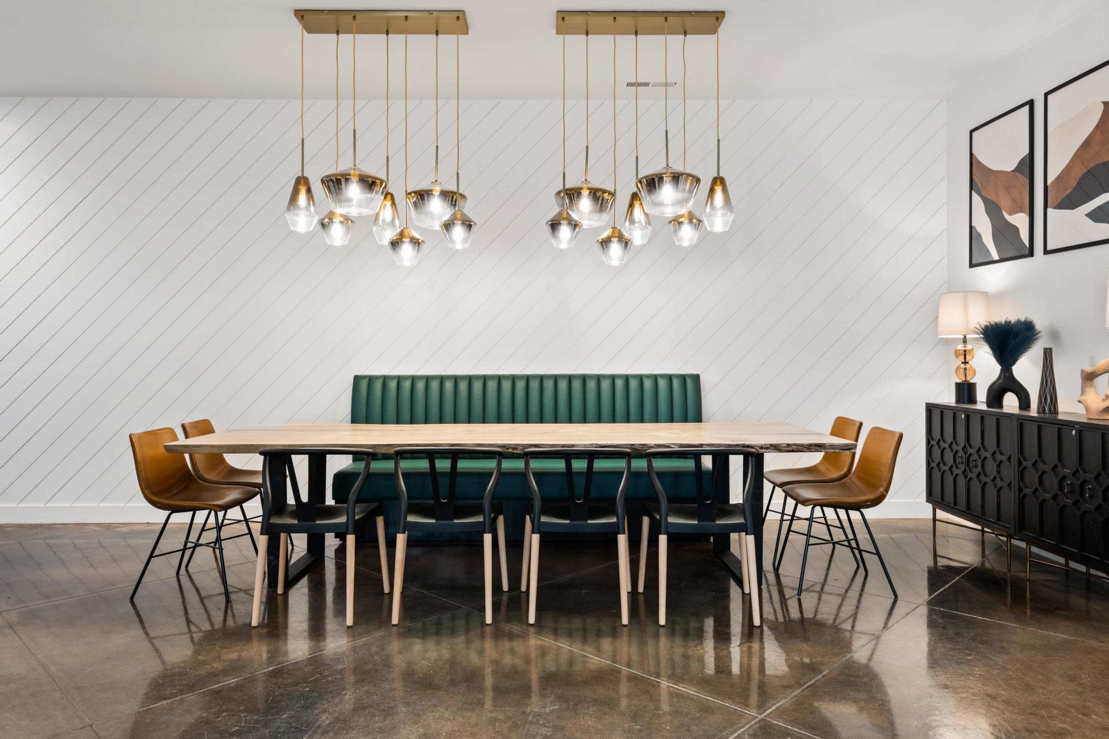 A long dining table with a green upholstered bench is positioned against a white striped wall, surrounded by six wooden chairs and illuminated by pendant lights above.
