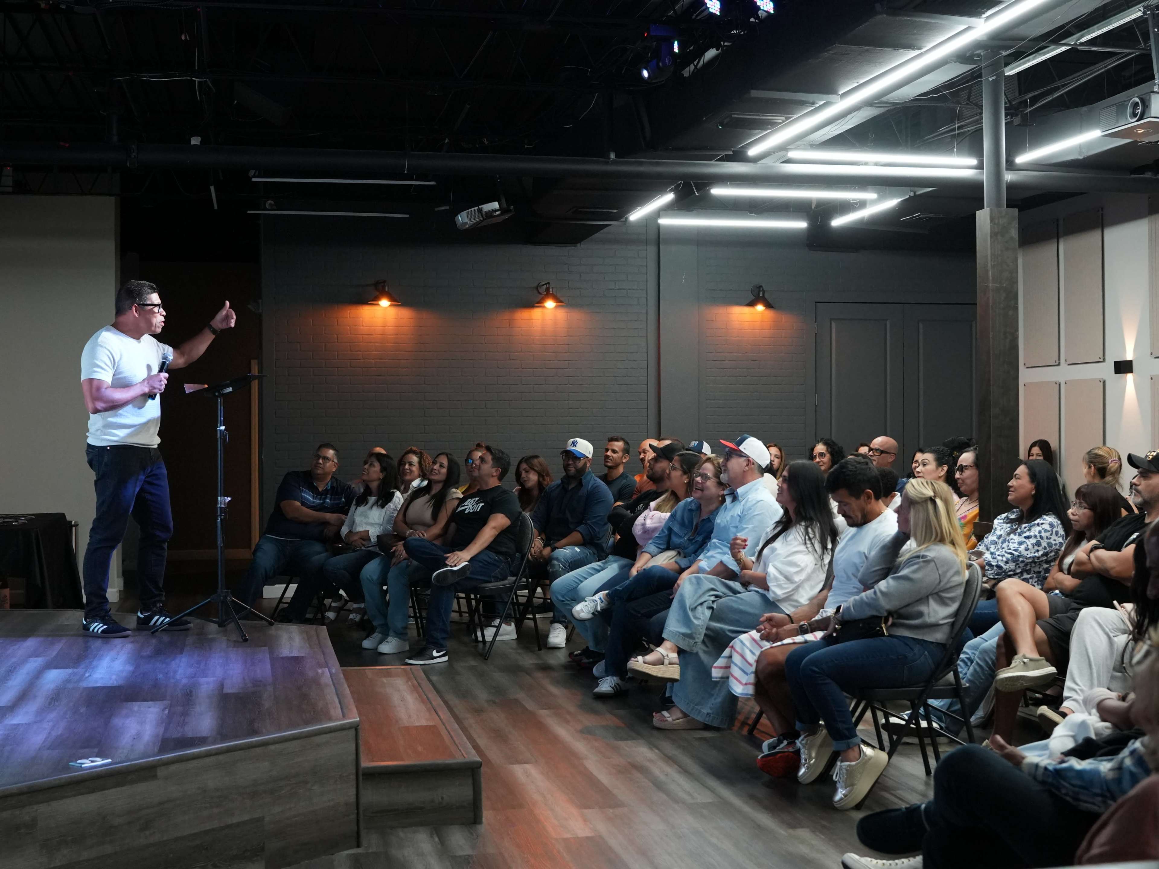 A speaker stands on a stage in front of an audience seated in chairs, engaged in a presentation within a well-lit venue.