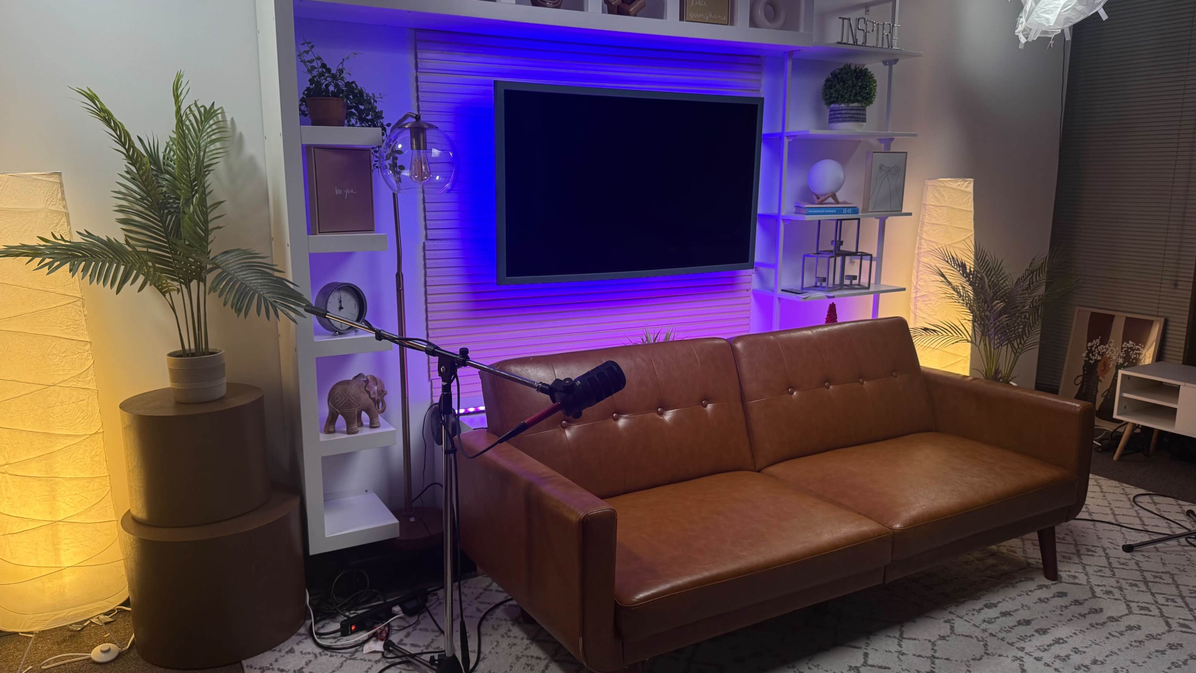 A modern living room features a brown leather sofa, a large wall-mounted television, and shelves with decorative items illuminated by colored lights.