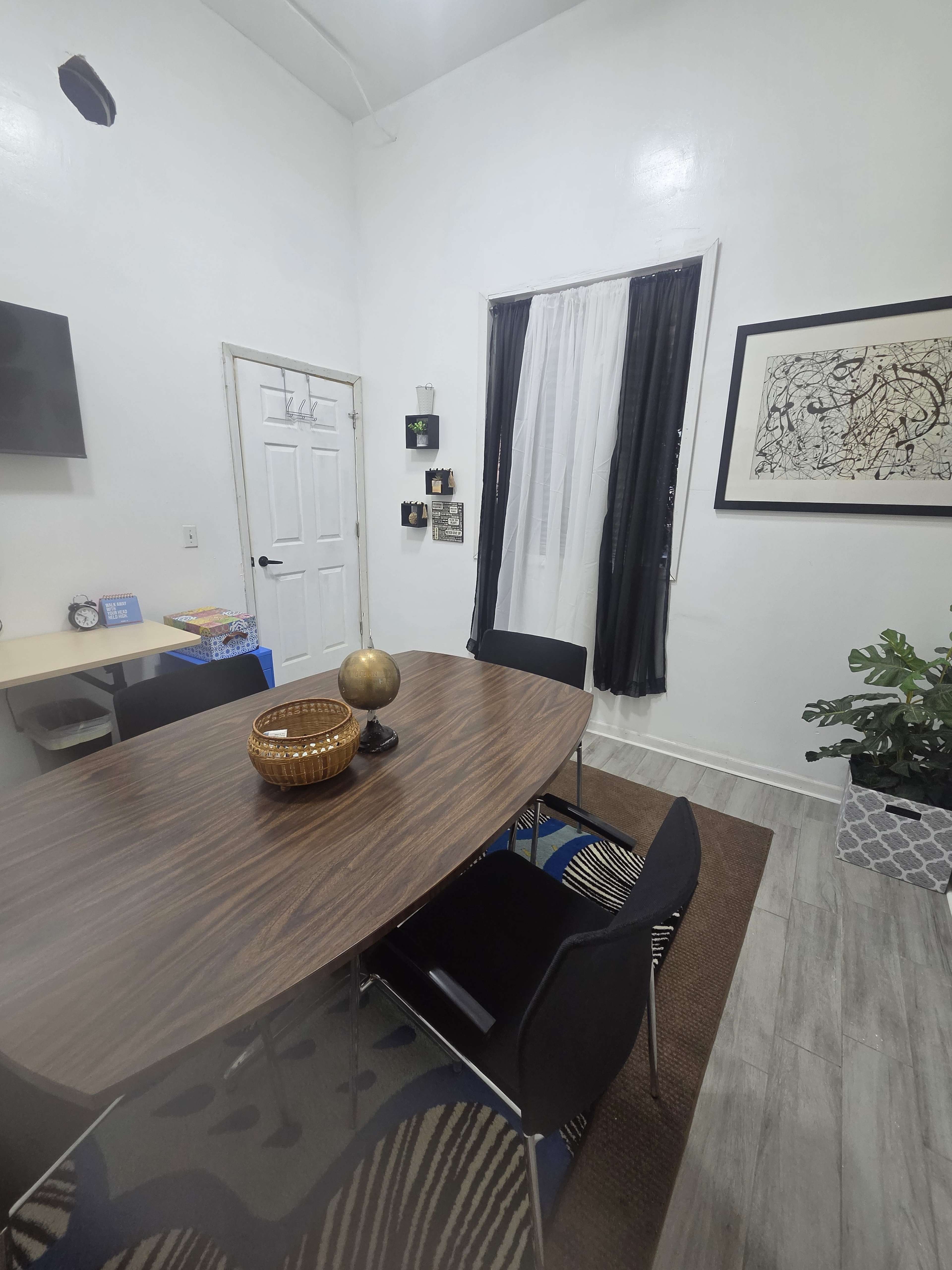 The image shows a small dining area with a wooden table, black chairs, a decorative bowl, and a door leading to another room.