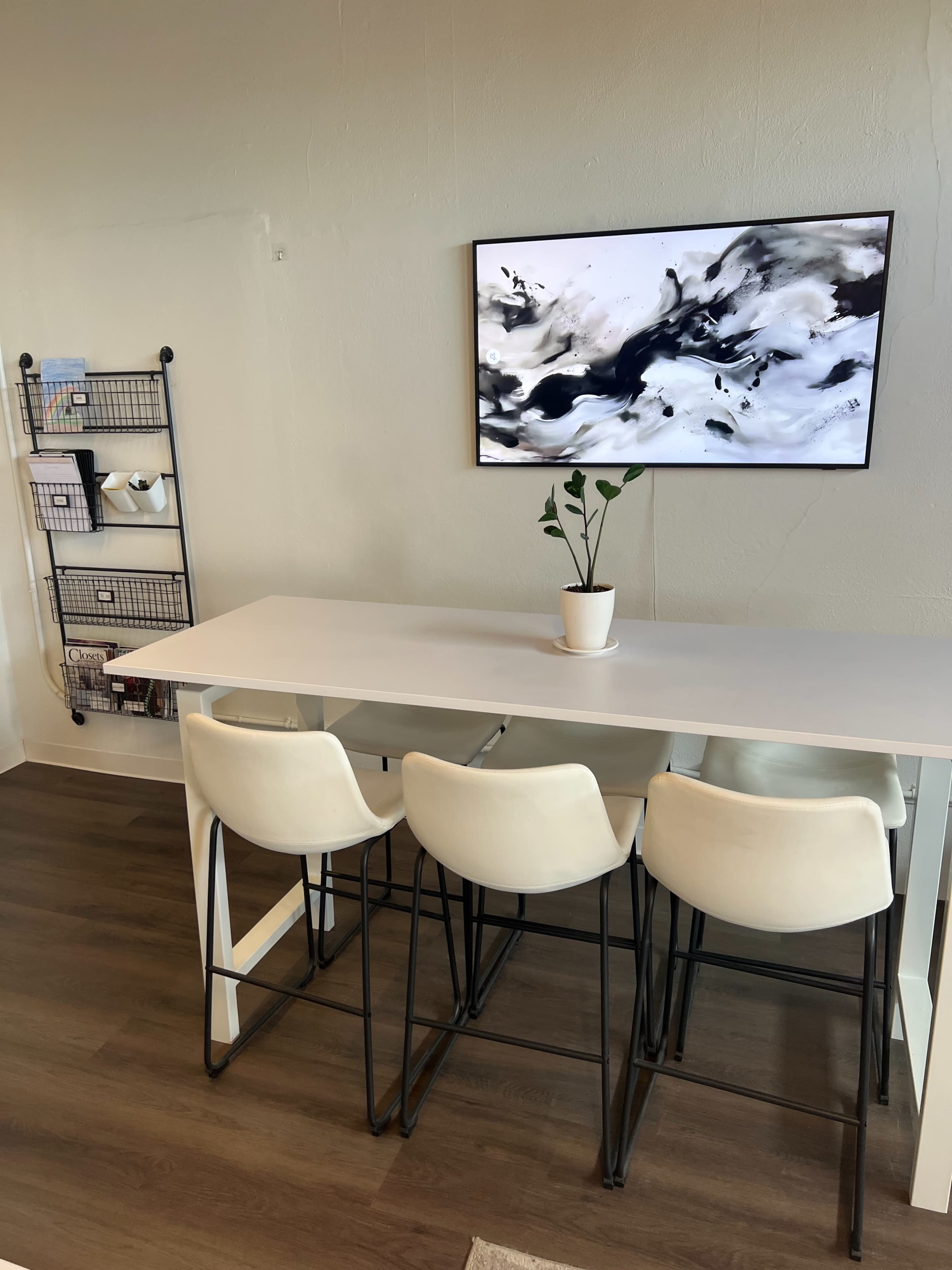 A minimalist dining area features a long white table with four white stools, a plant in a pot, and an abstract artwork on the wall behind.
