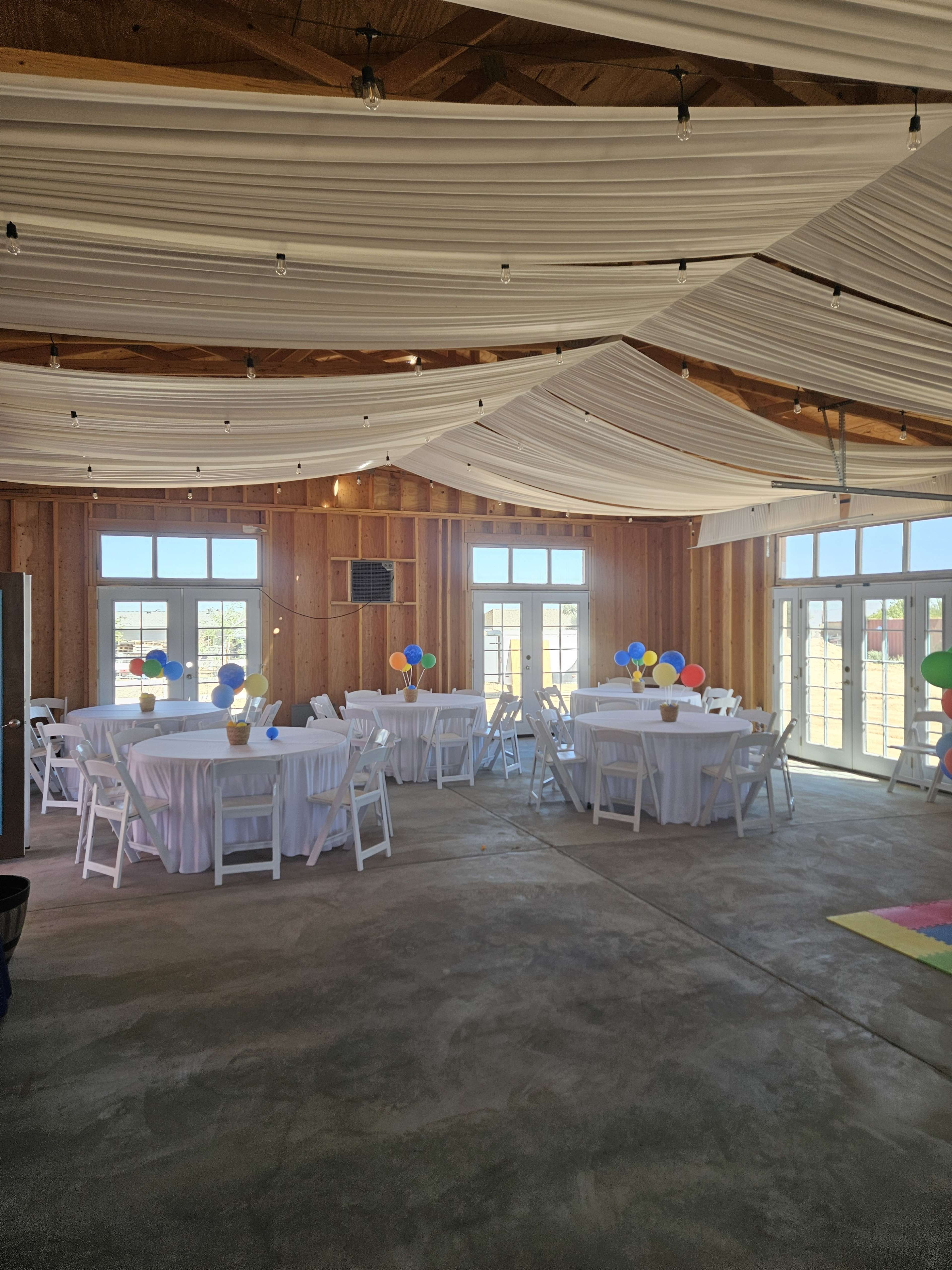 The image shows a spacious event venue with tables covered in white tablecloths, colorful balloons, and draped white fabric overhead.