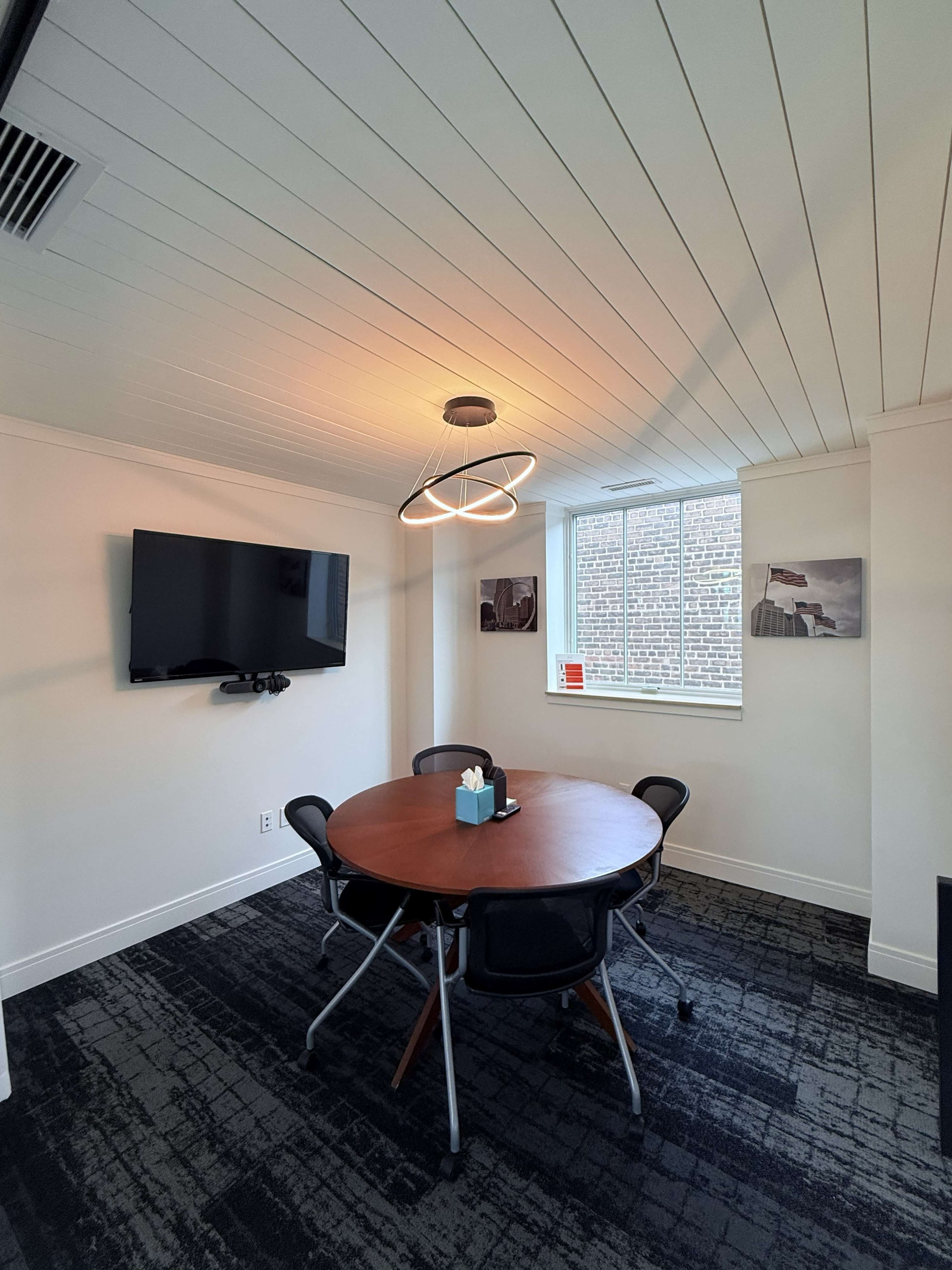 The image shows a modern conference room with a round wooden table, four black chairs, a wall-mounted TV, and a window providing natural light.