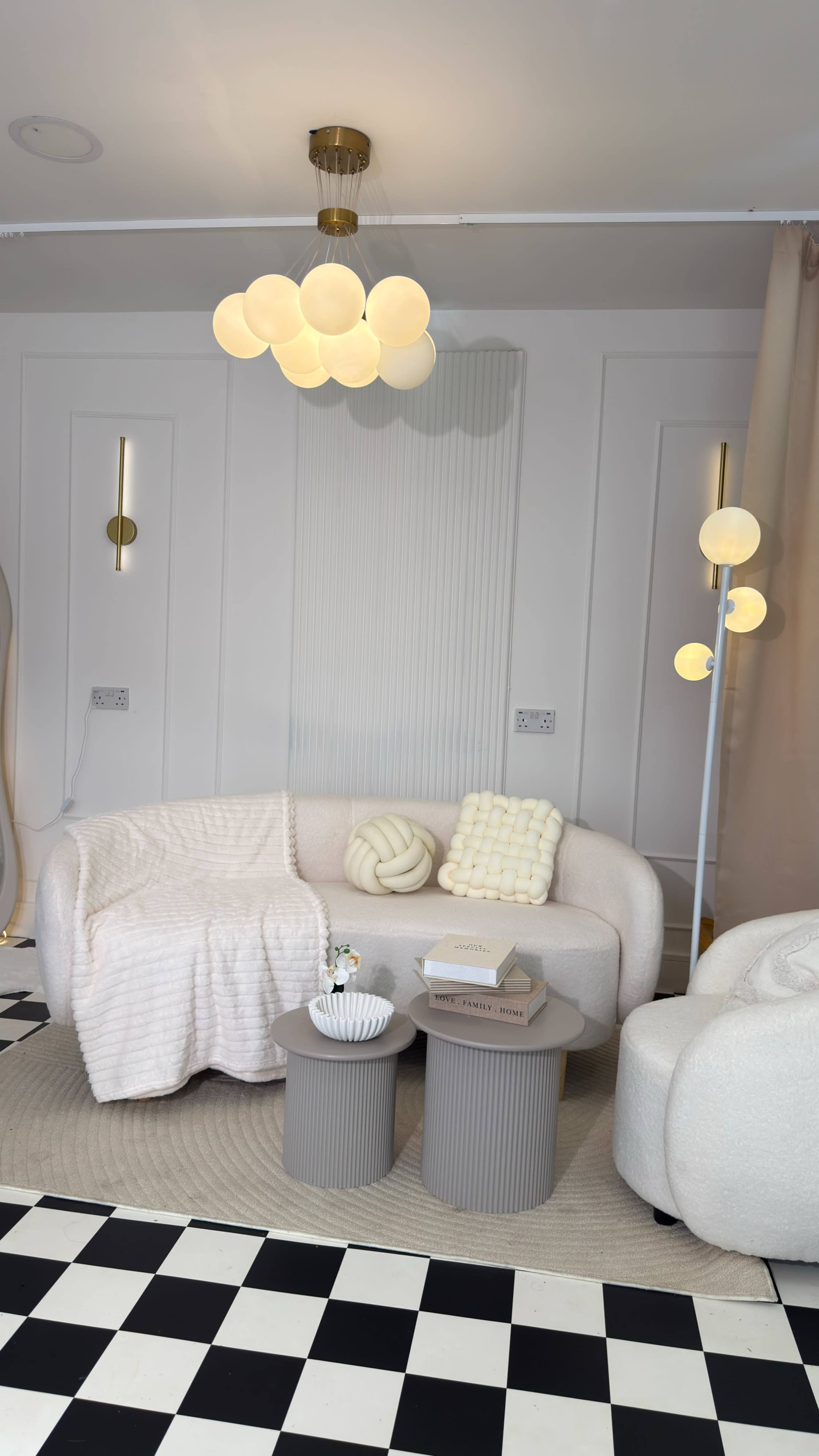 The image shows a modern living room with a white curved sofa, a pair of round side tables, and a unique ceiling light fixture, all set against a checkered floor and light-colored walls.
