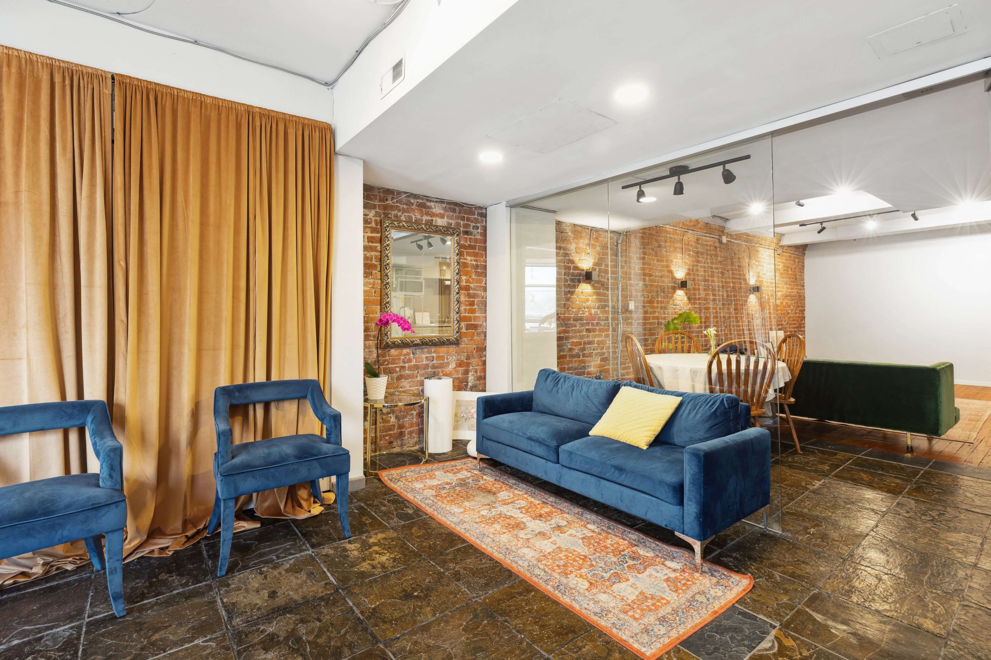 A spacious living area featuring a blue velvet sofa, two matching chairs, a patterned rug, and exposed brick walls, with a glass partition leading to another room.