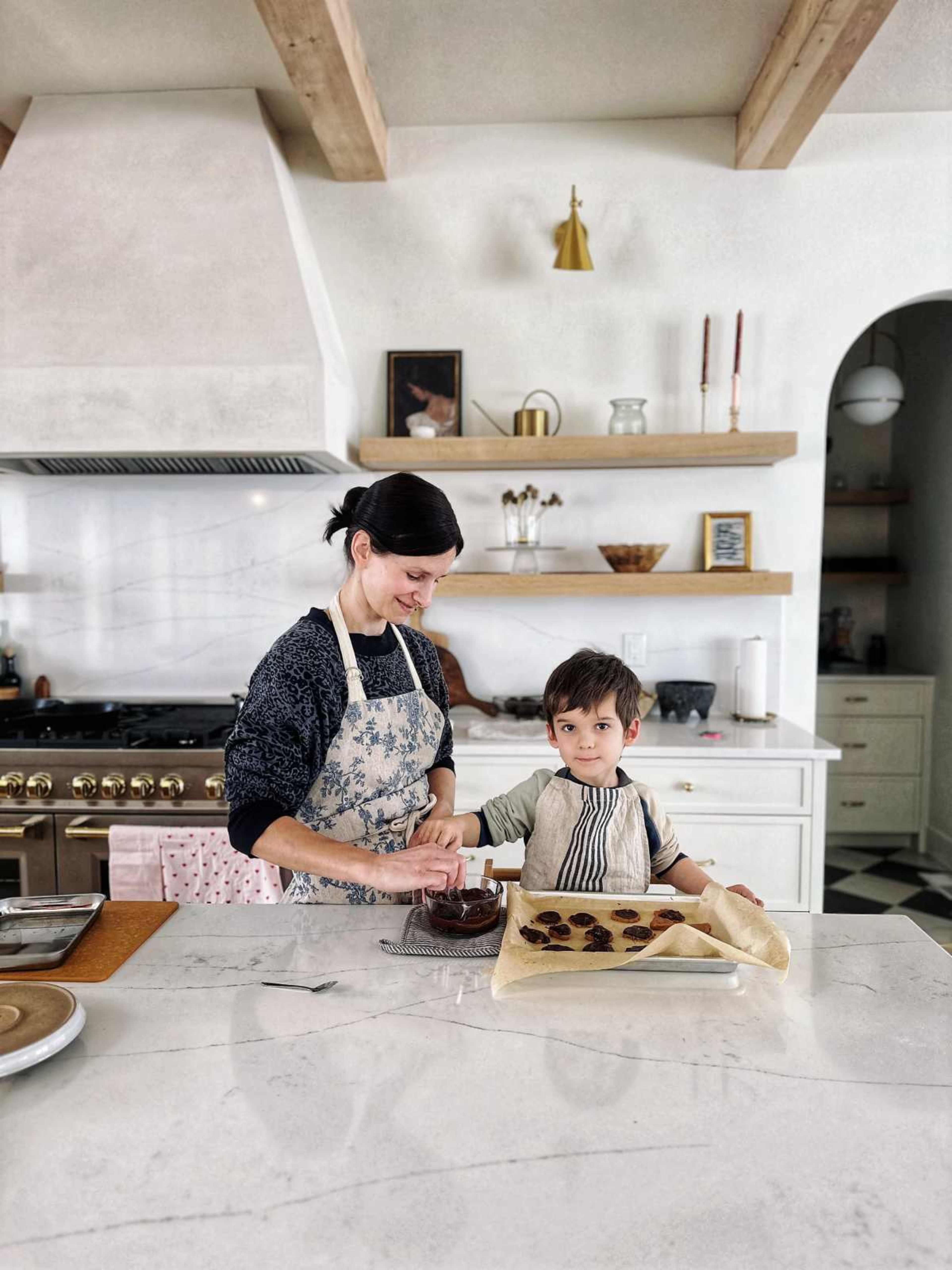 A woman and a young boy are baking together in a modern kitchen, with the boy helping to prepare cookie dough on a marble countertop.