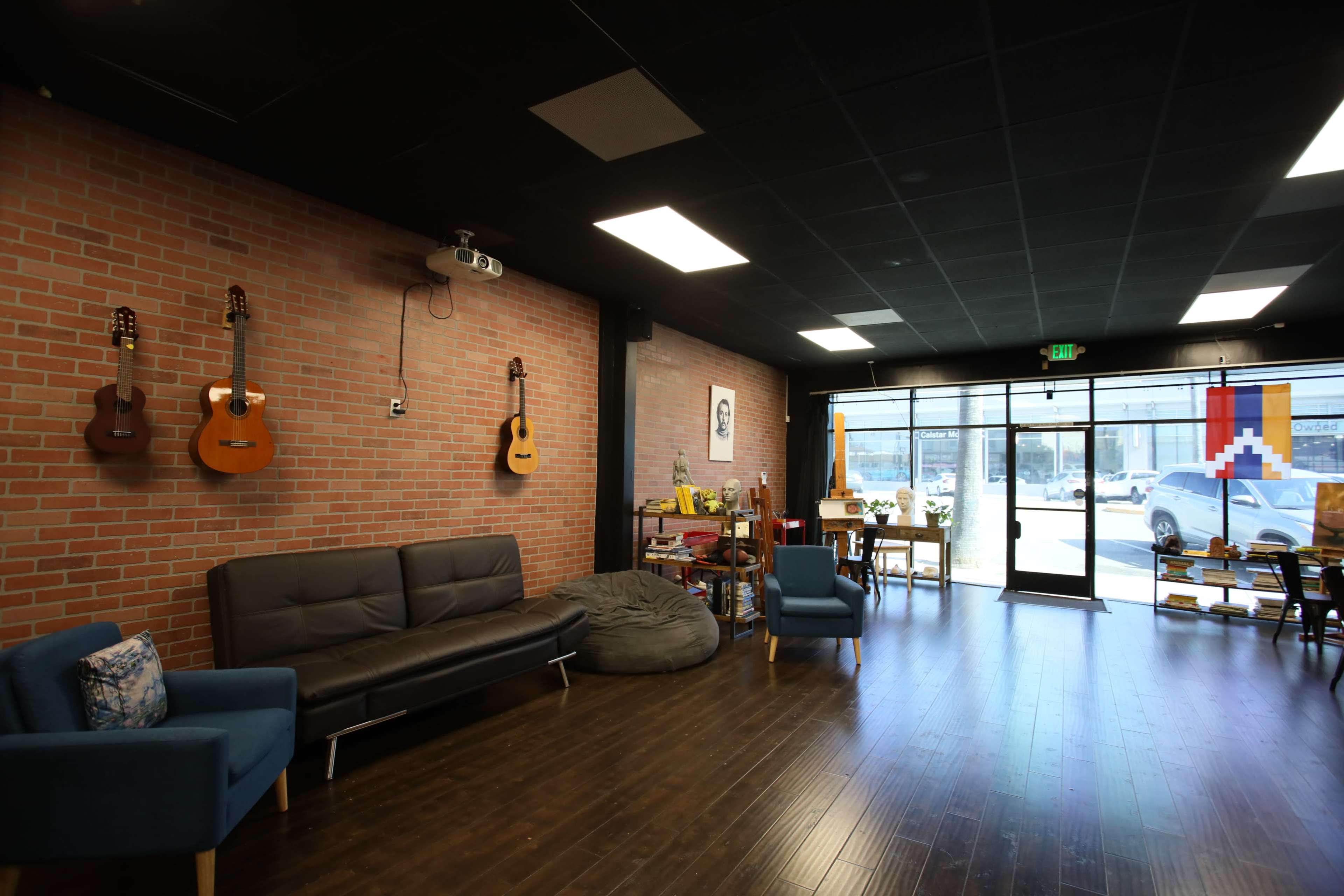 The interior of a cozy space features a brick wall with guitars hung up, a dark sofa, a bean bag, and tables in front of a glass entrance.