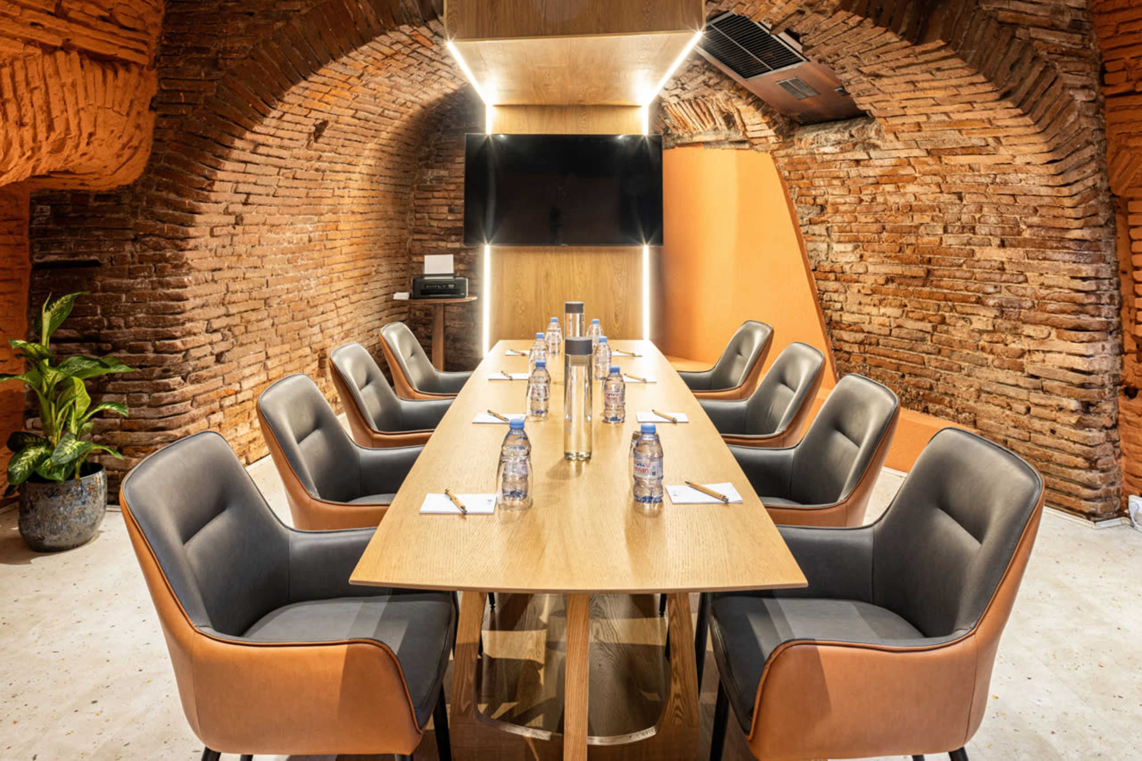 A modern conference room features a long table surrounded by eight ergonomic chairs, with a large screen mounted on a brick wall.