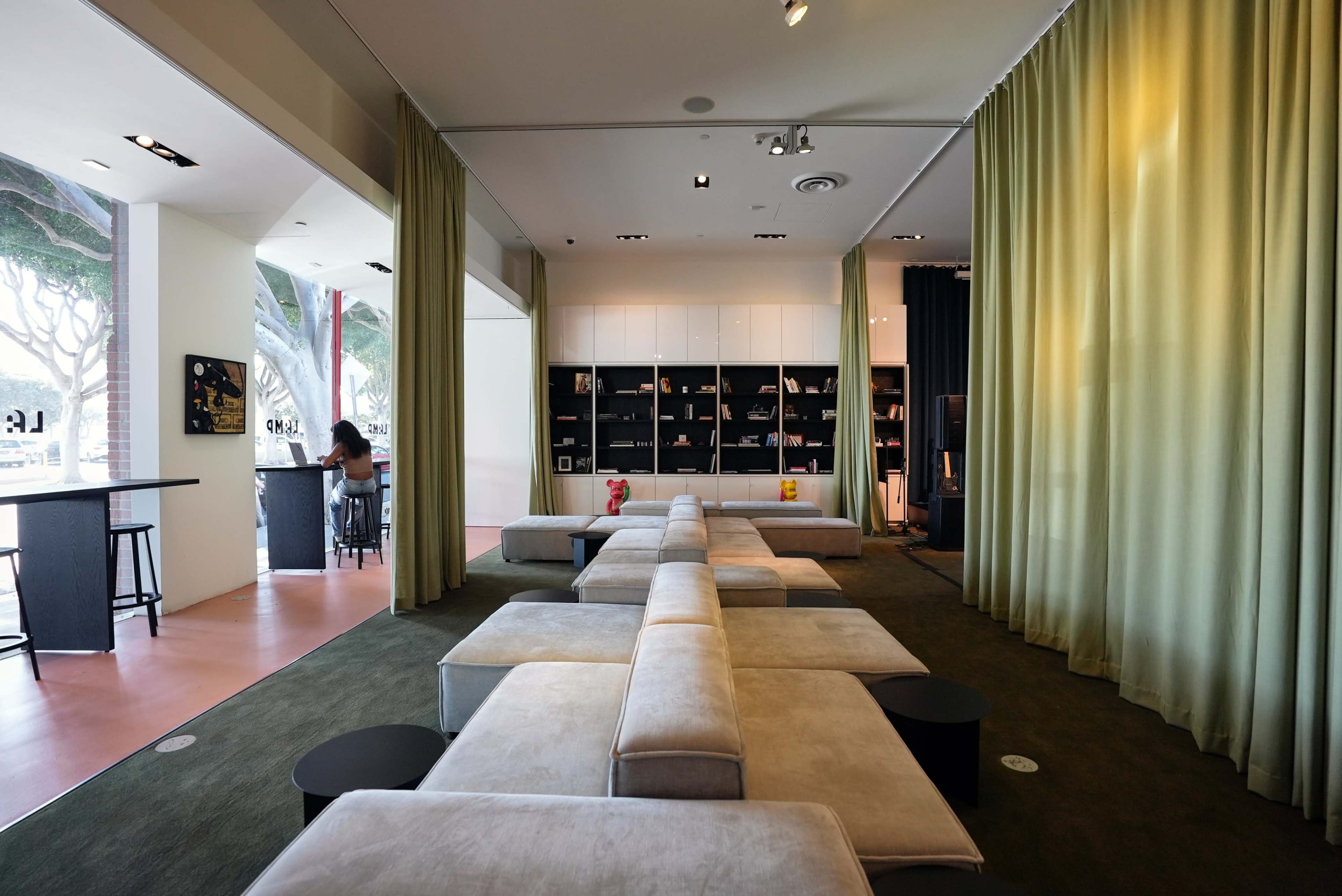 The image shows a modern interior space with a seating area featuring low, rectangular sofas, a shelf filled with books and decorative items, and a bar-style table in the background.